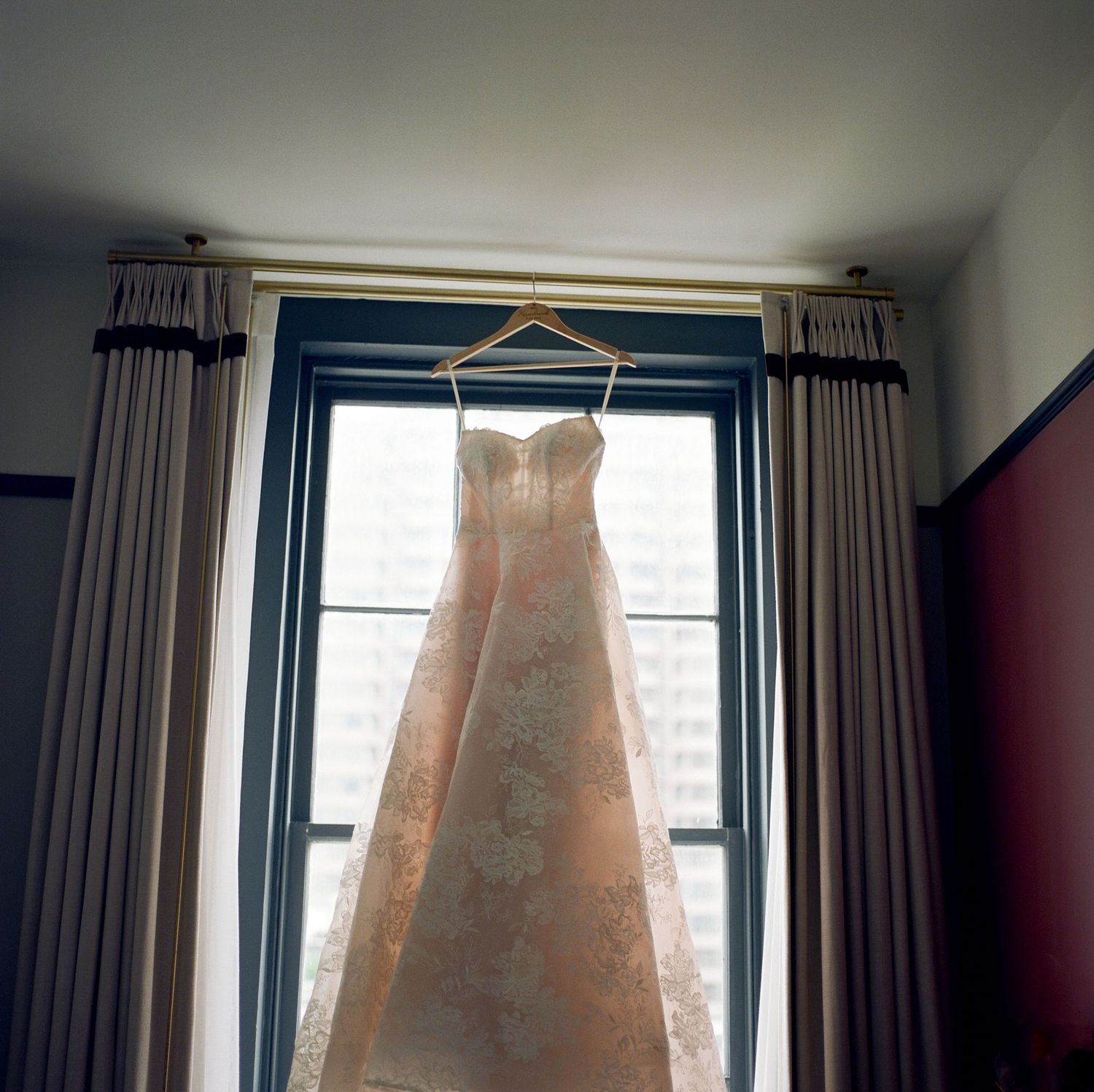 Wedding dress with intricate lace details hangs in window with natural light streaming through.
