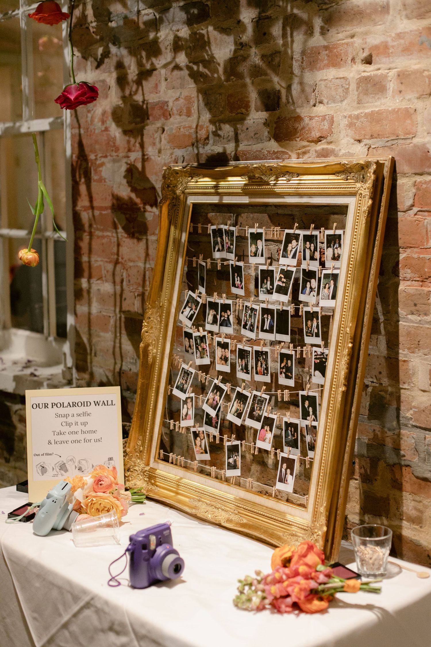 Wedding photo display with gold frames and polaroid pictures arranged on a white tablecloth against brick walls.