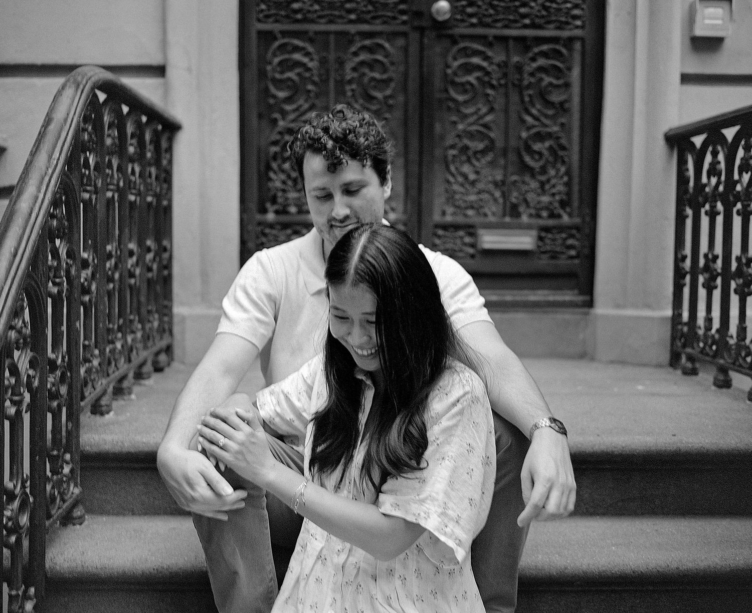 Black and white photo of a couple sitting on brownstone steps, man embracing woman from behind near ornate door.