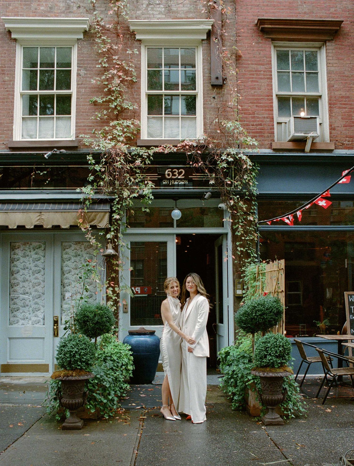 A charming brick storefront with climbing vines and potted plants frames two people in white outfits.