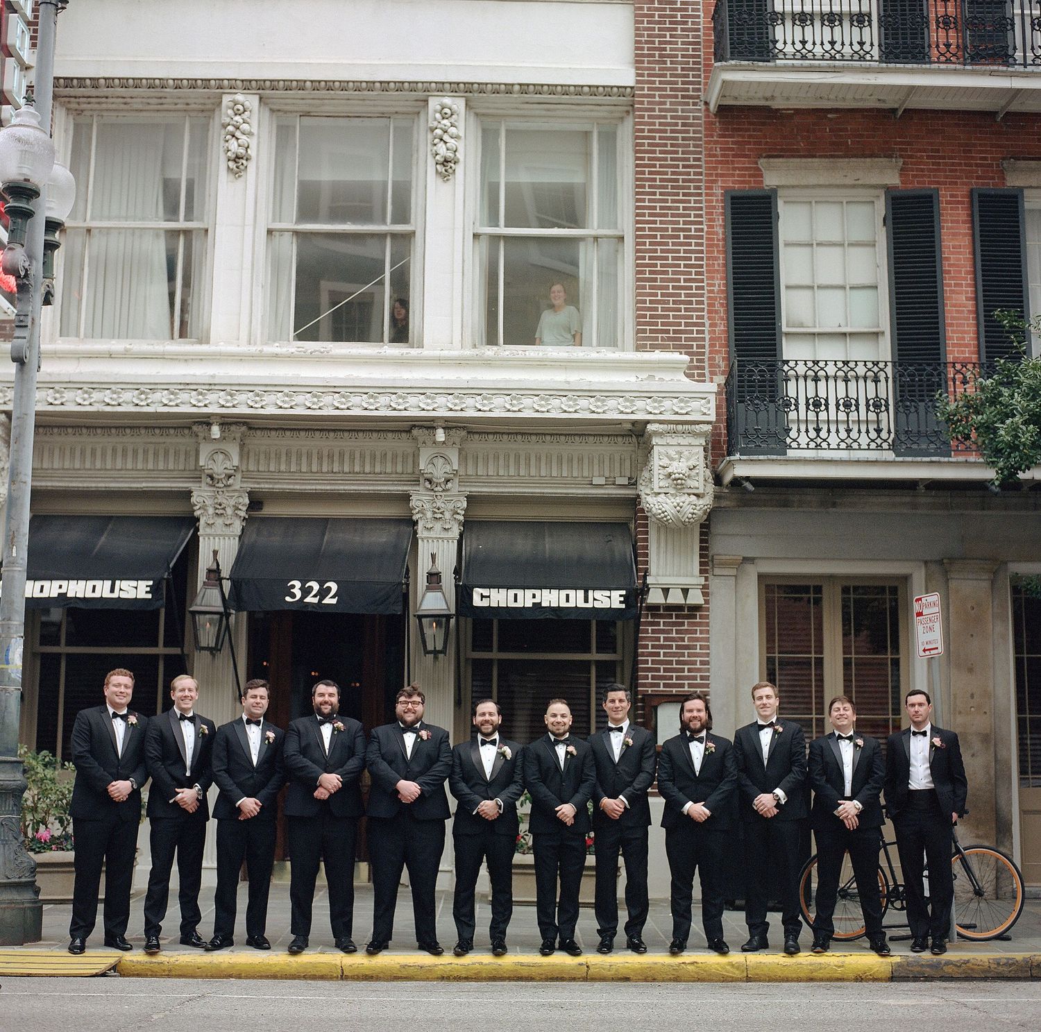 Wedding party groomsmen in formal attire stand in line against urban brick building backdrop.