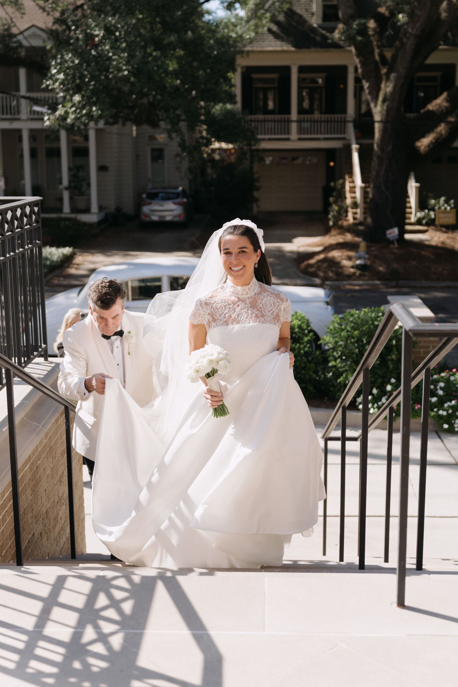 A bride in a lace gown ascends outdoor stairs while holding her dress.