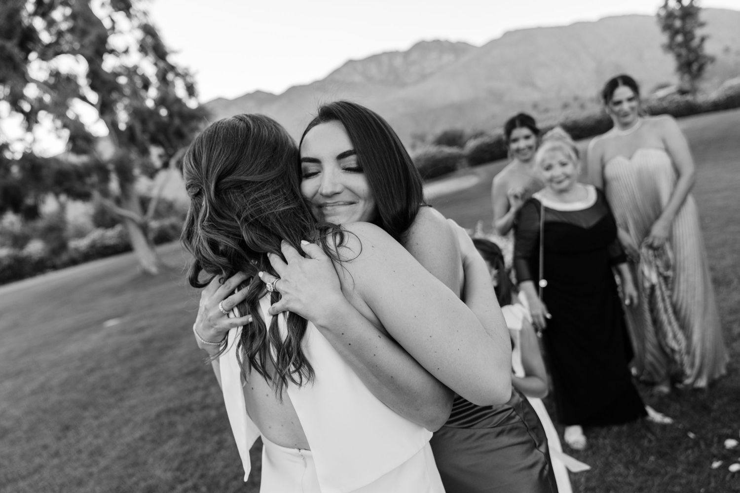 Two wedding guests share an emotional embrace during an outdoor celebration with mountains in the background.