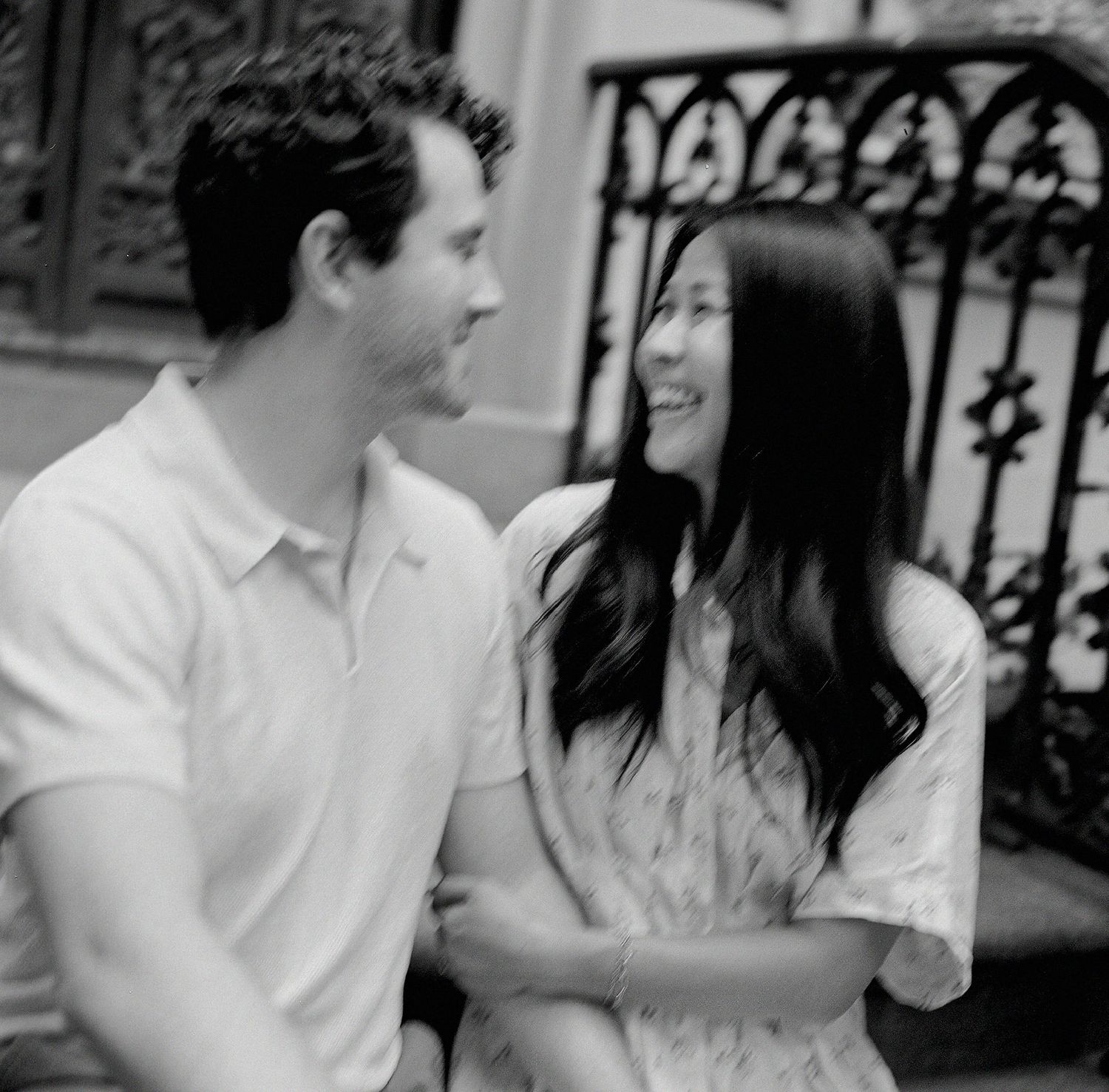Black and white photo of a smiling couple sharing a tender moment outdoors near an ornate iron railing.