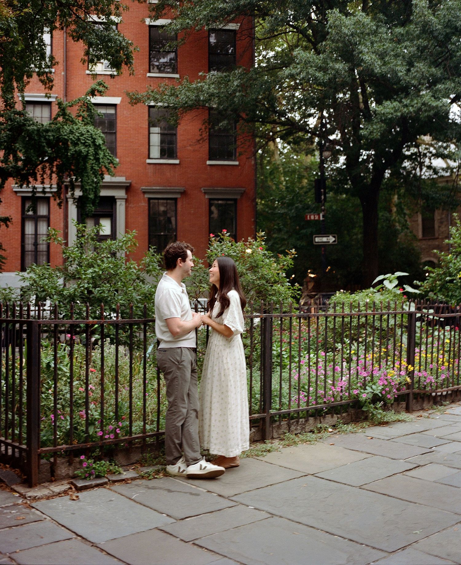 Couple standing together by iron fence with garden and red brick building in urban neighborhood.