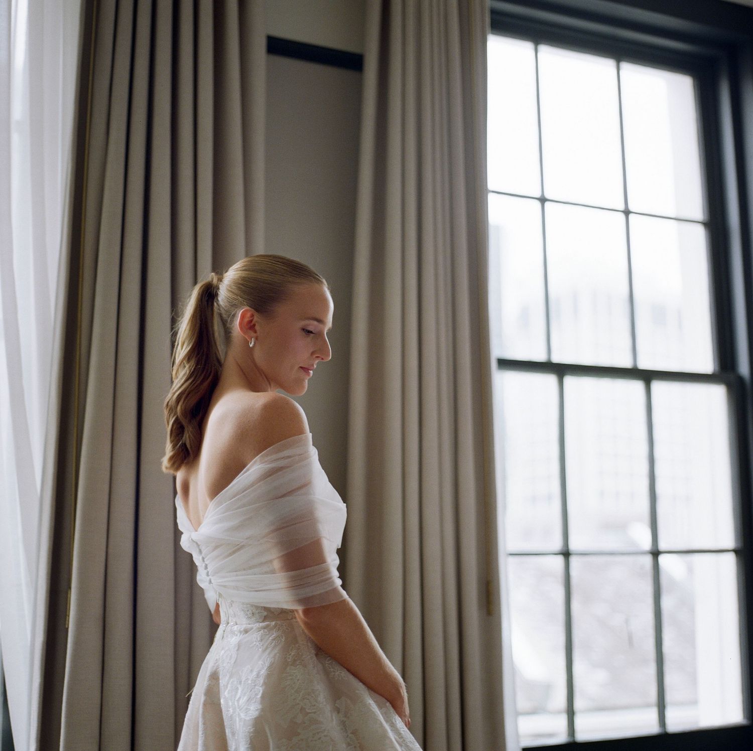 Bride in white dress poses by window with natural light streaming in.