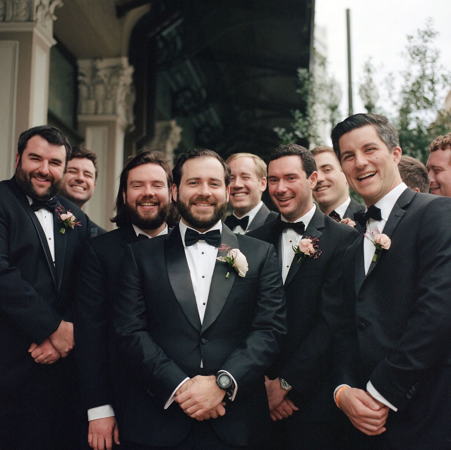 Groomsmen in black tuxedos share laughs during outdoor wedding photo session.