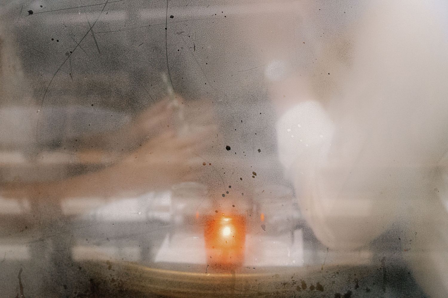 Blurry ethereal image of a glowing orange candle flame on a table, seen through foggy glass with dark spots.
