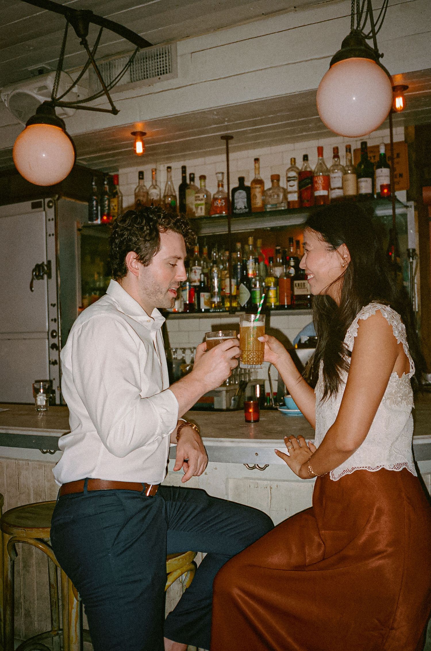 Couple toasting cocktails at a bar with bottle-lined shelves, sharing a romantic moment under warm globe lights.
