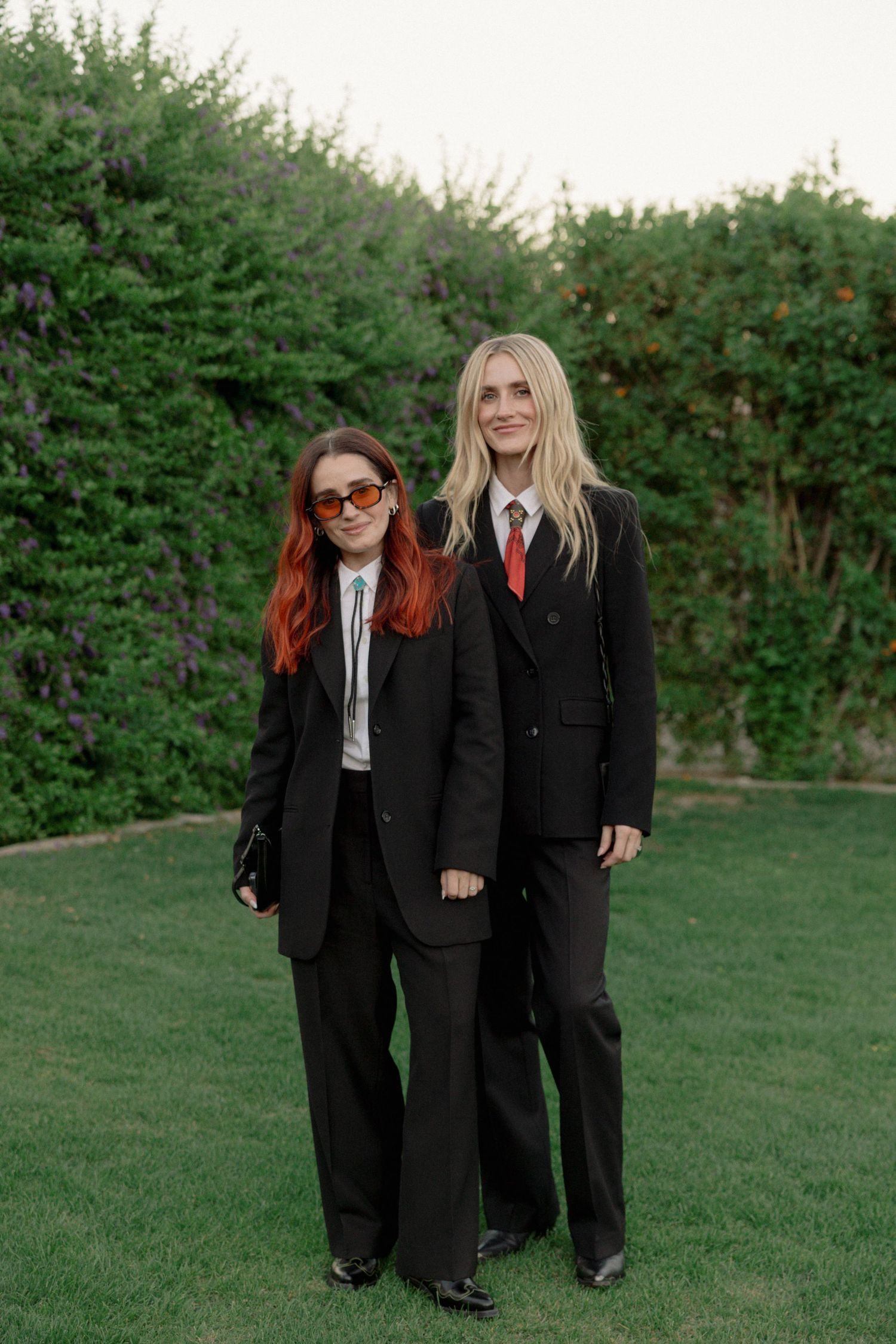 Two people in matching black suits and red ties stand together on a grassy lawn with green hedges in the background.