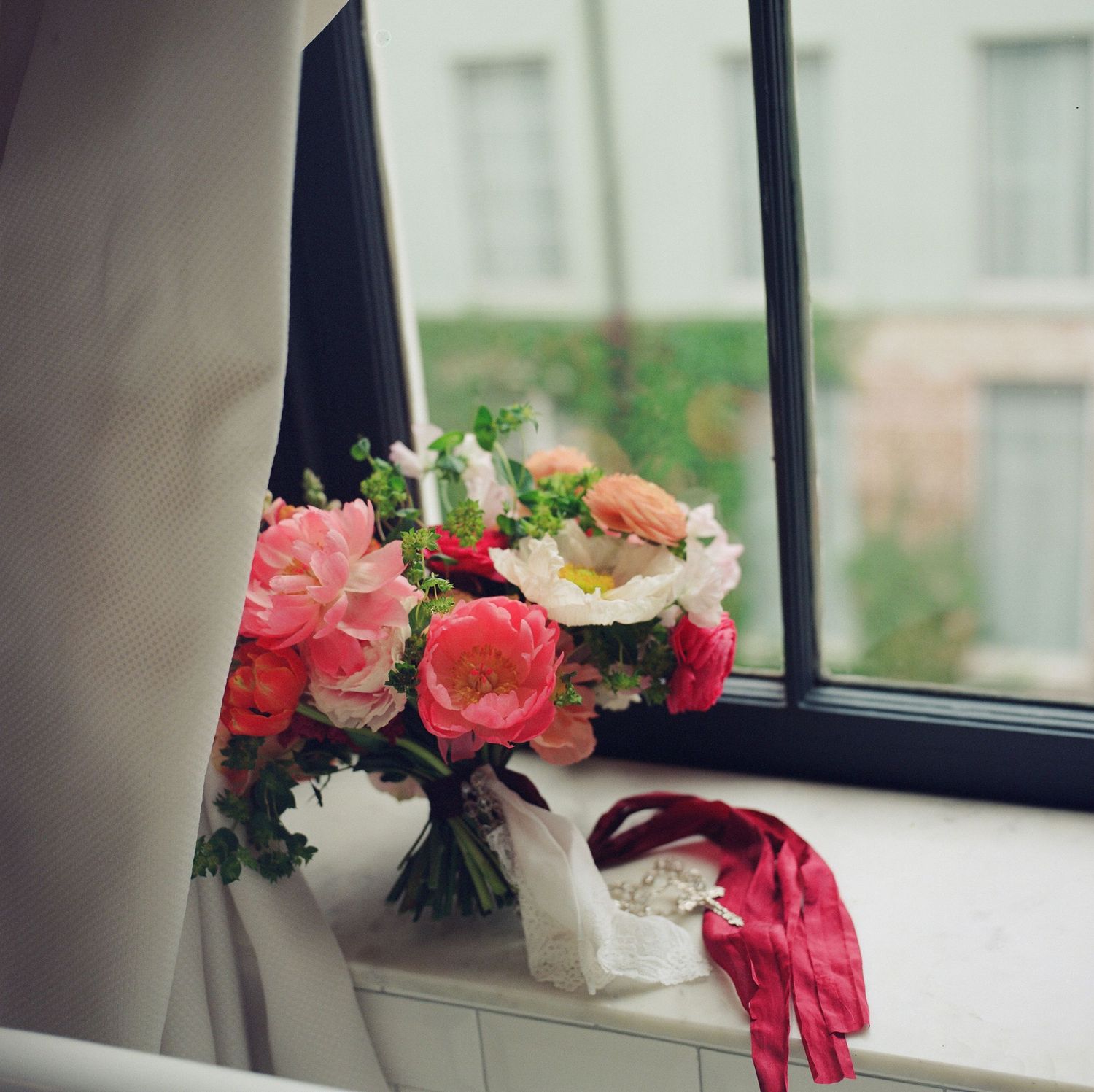 Pink and coral floral bouquet arranged near a window with wedding accessories.
