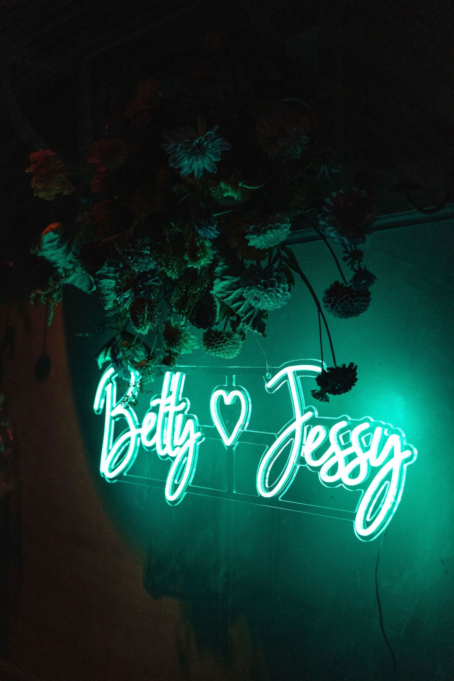 Turquoise neon sign with cursive text illuminated against dark background with hanging foliage.