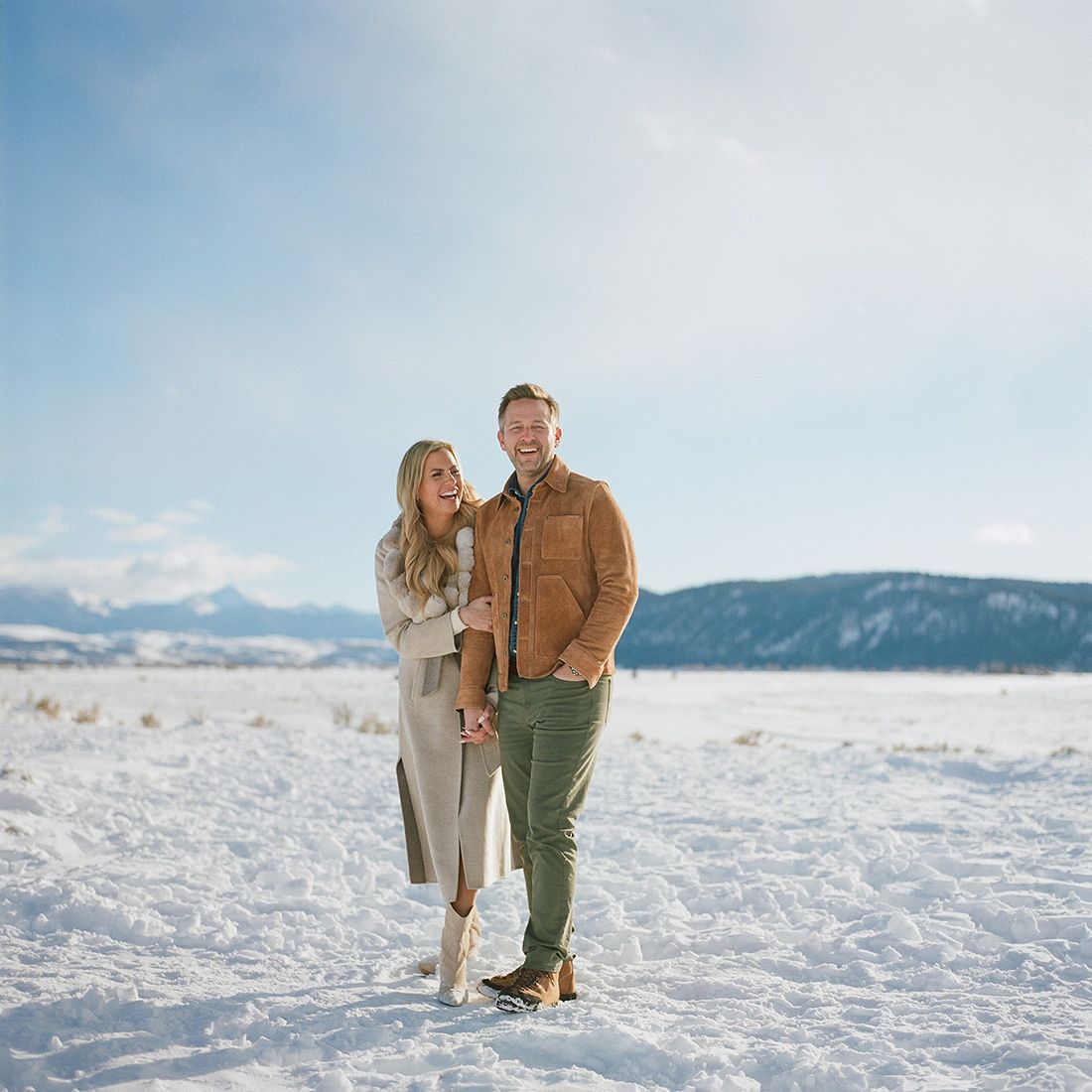 A couple poses together on a snowy landscape with mountains in the distance.