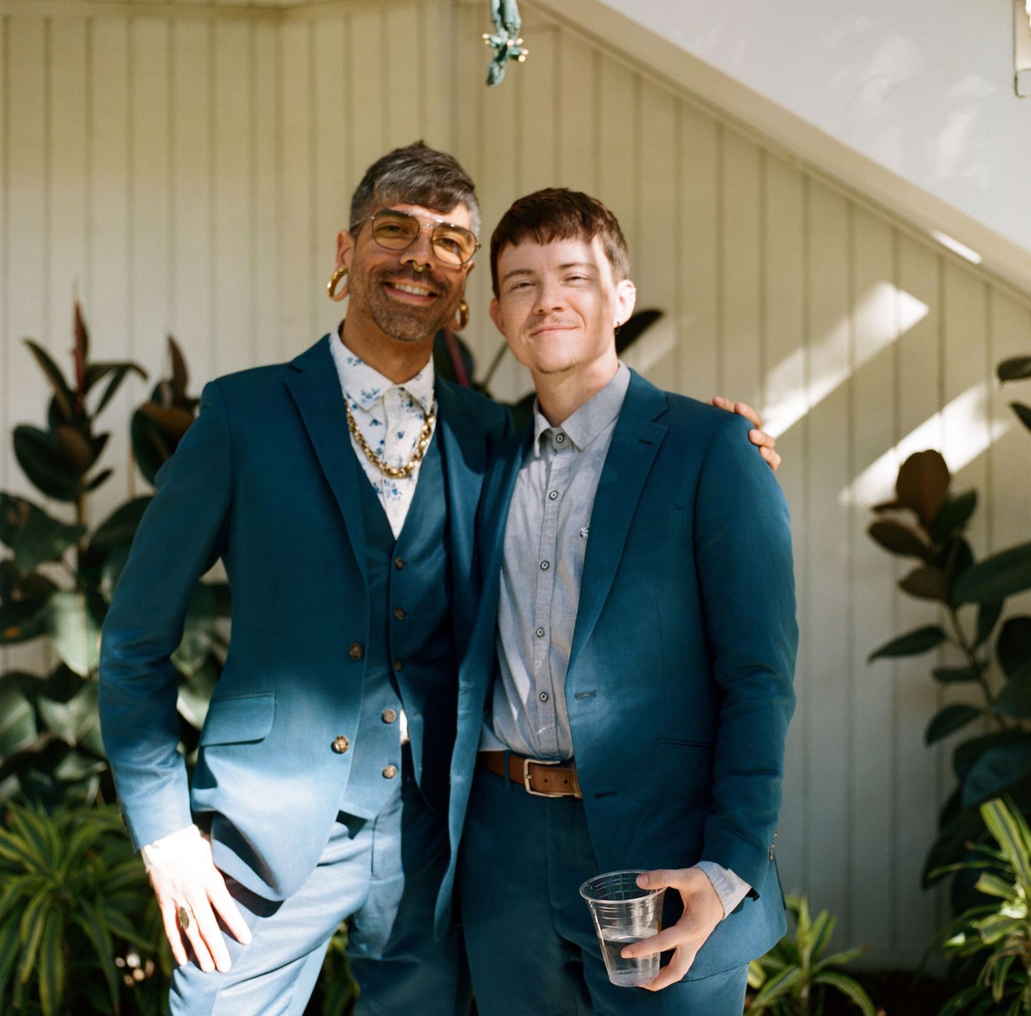 Two wedding guests in navy suits pose together at an outdoor venue.