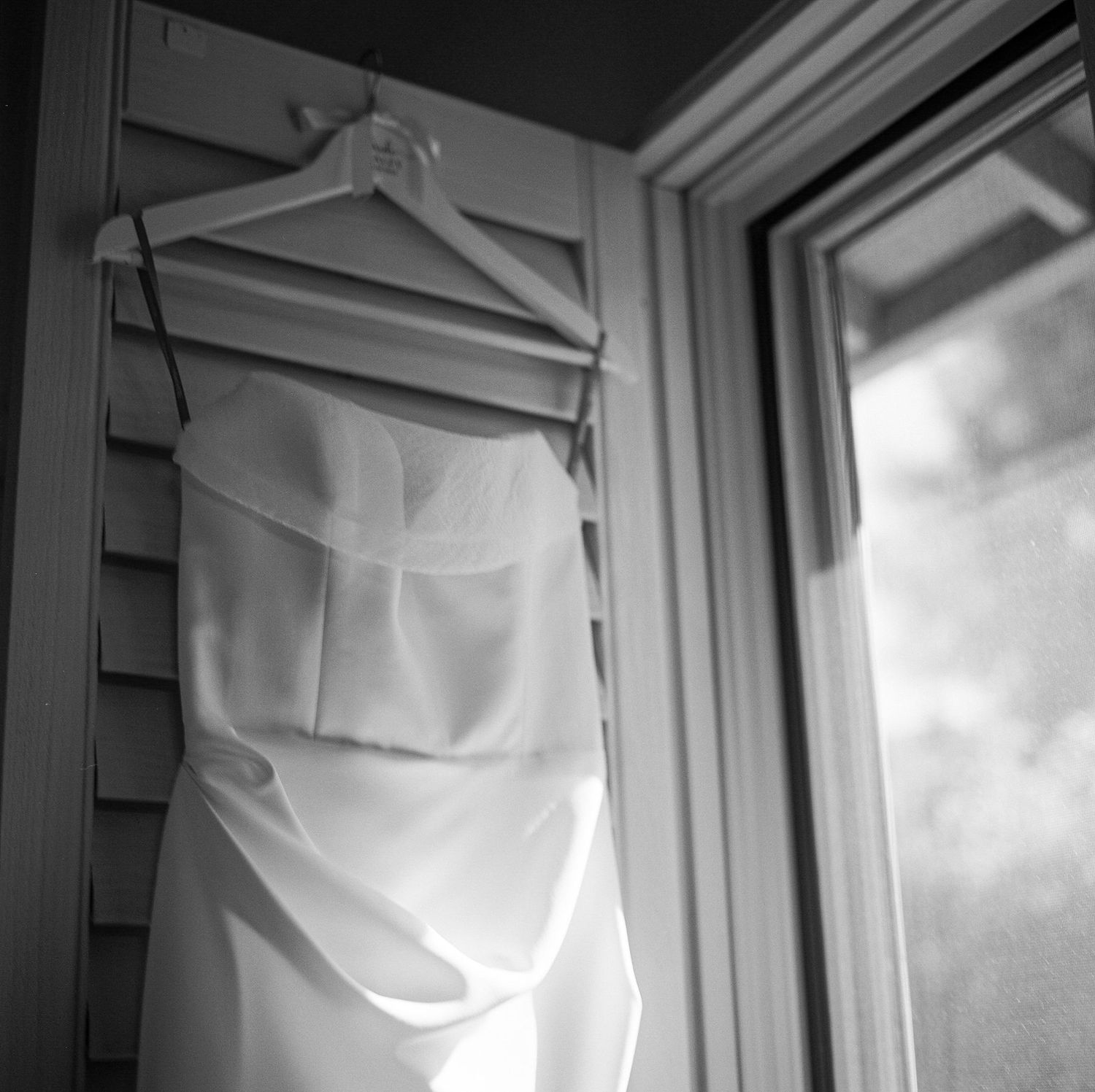 Black and white photo of a white wedding dress hanging by a window on a wooden hanger.