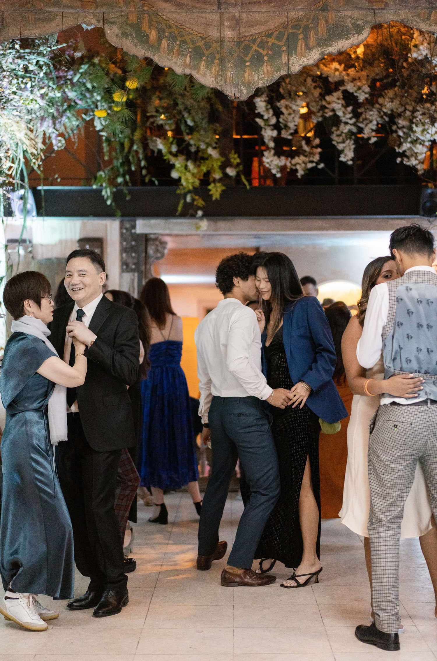 Wedding guests dancing beneath hanging floral decorations at an indoor reception.