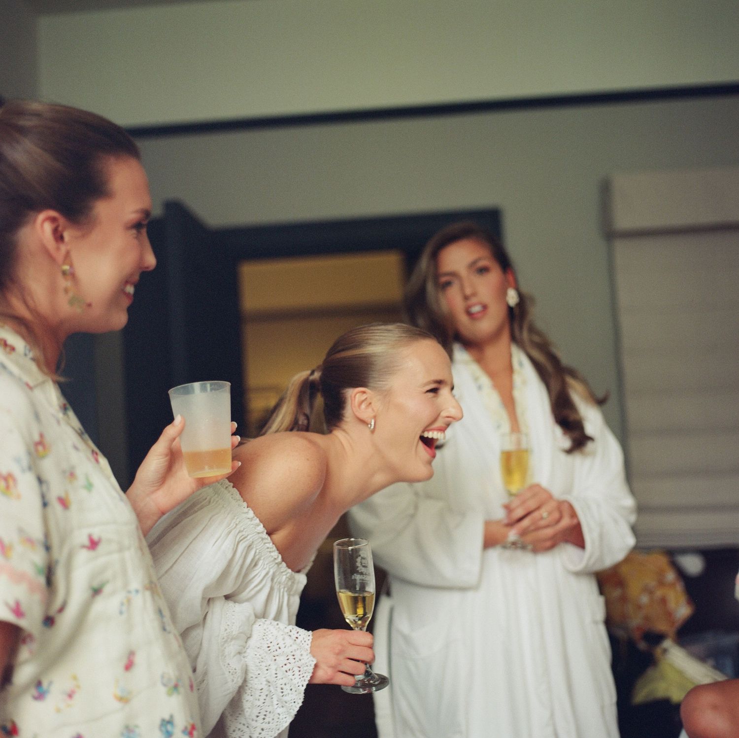 Bridesmaids in white robes share champagne and laughter during wedding preparations.
