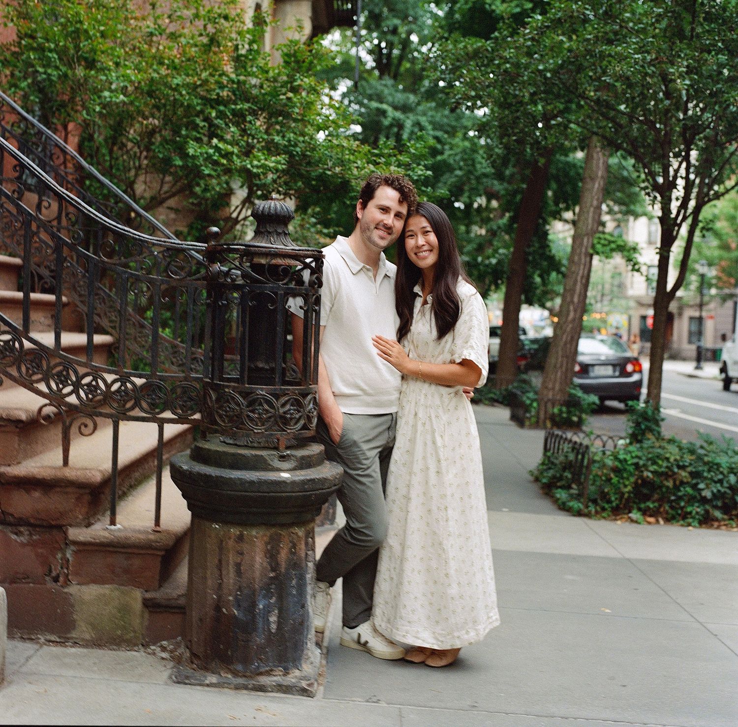 Couple smiling together on a tree-lined NYC sidewalk near ornate iron staircase railing.
