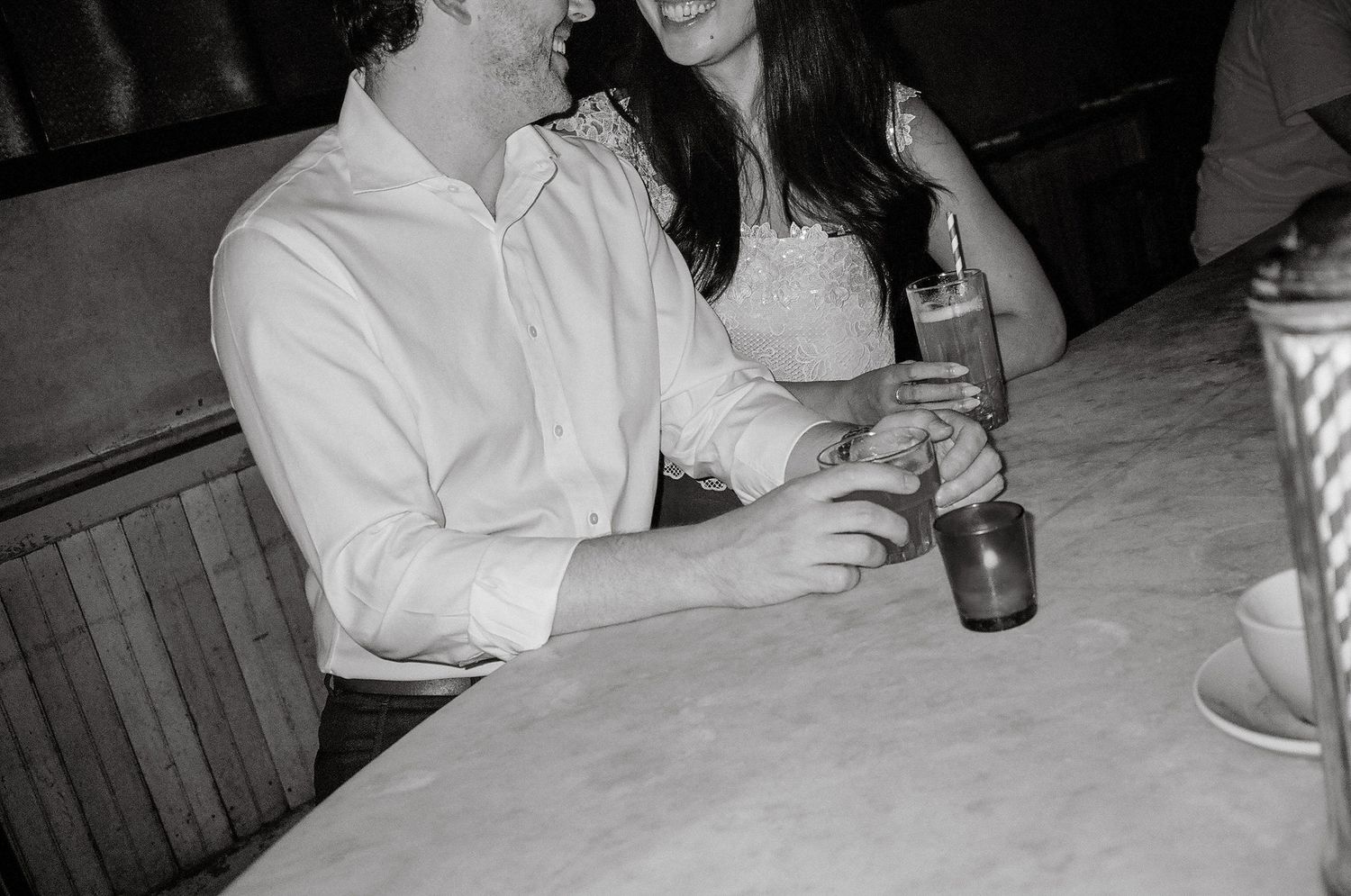 Black and white photo of a couple sitting closely at a marble table, sharing drinks in a cozy restaurant booth.