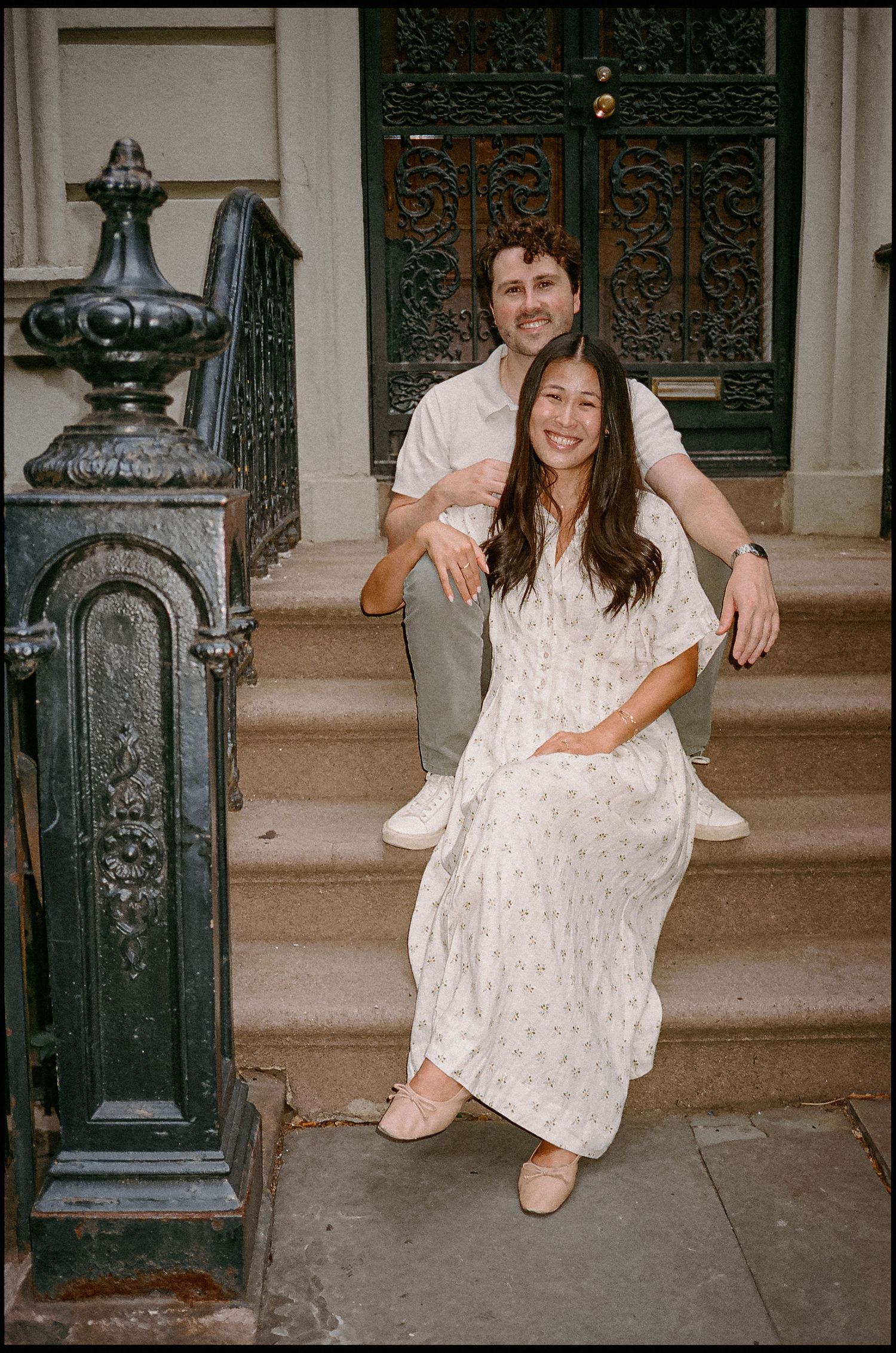 A smiling couple sits on brownstone steps, woman in floral dress, man in white shirt, urban NYC backdrop.