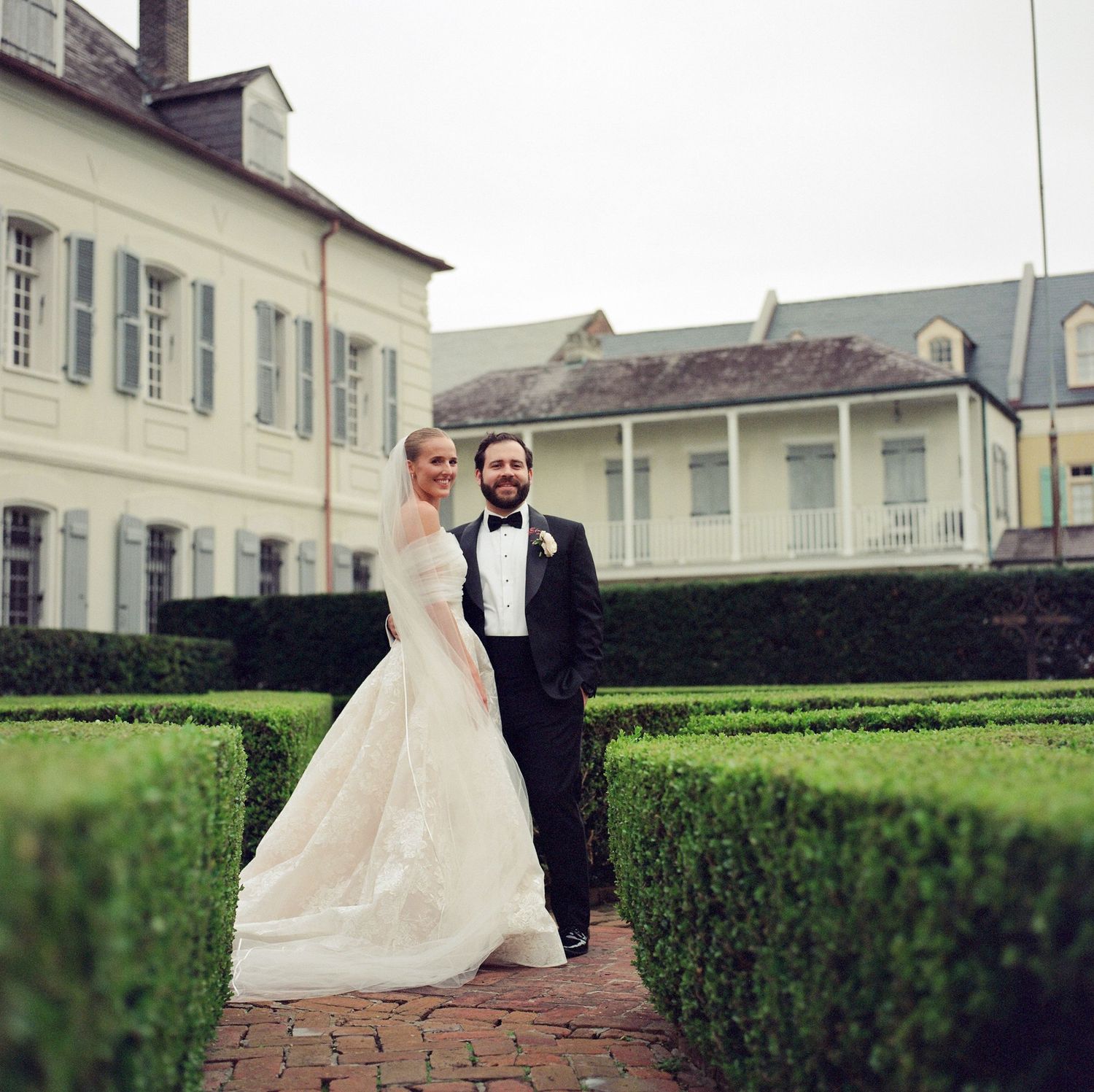 A couple walks along a brick pathway surrounded by perfectly manicured hedges at their wedding venue.