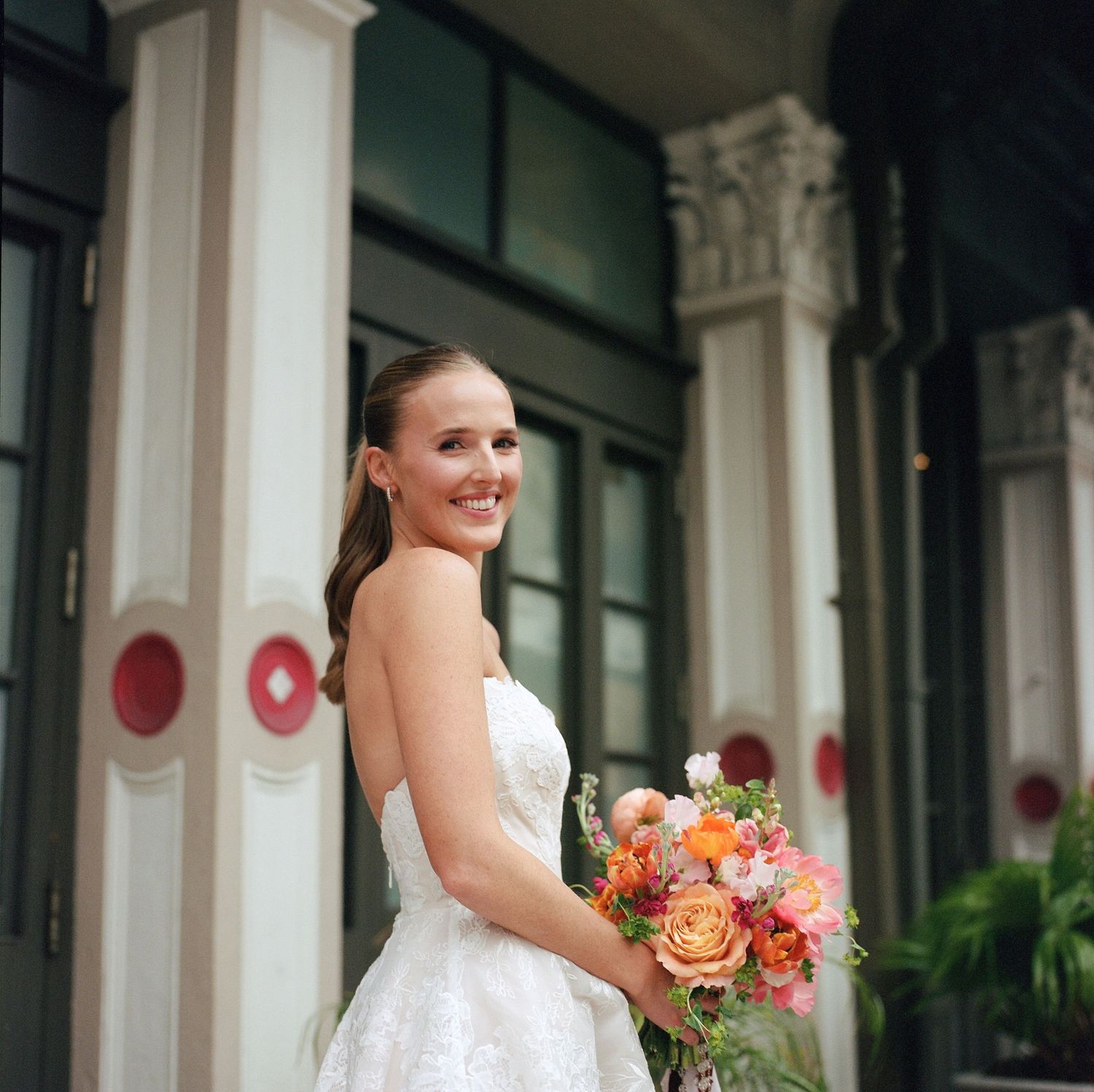 Bride holding vibrant orange and pink bouquet poses in front of storefront windows.