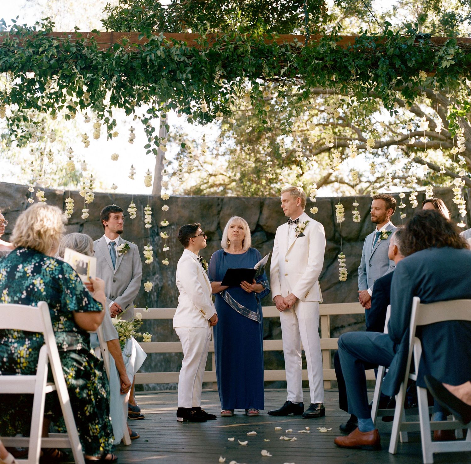 Wedding guests seated in white chairs on a wooden deck under flowering trees.