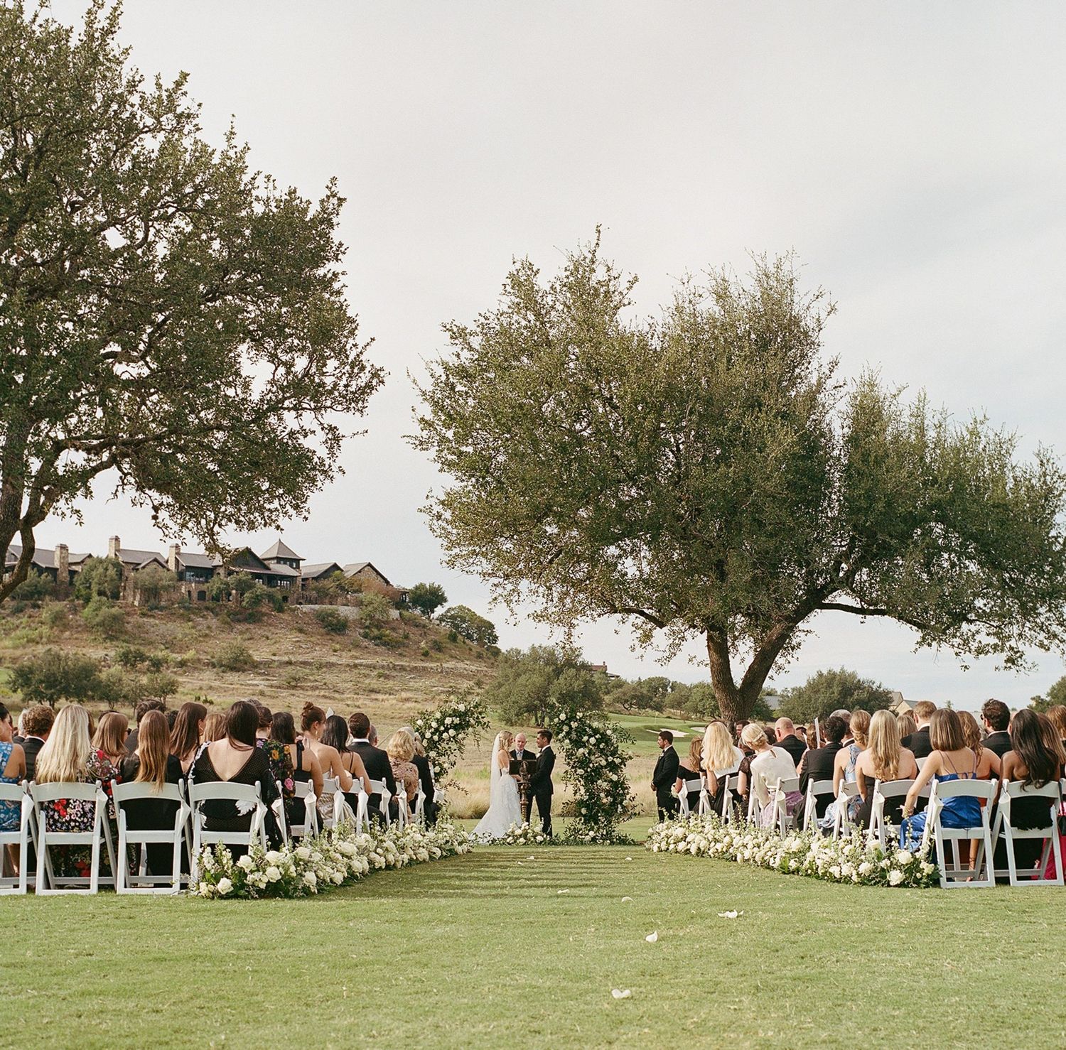 Outdoor wedding ceremony setup with white chairs arranged on a grassy hillside under trees.
