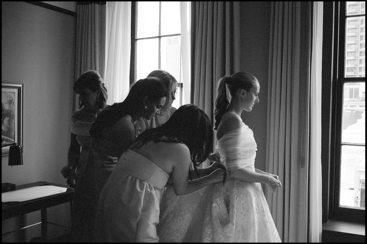 Black and white photo of bridesmaids helping bride prepare near window before wedding ceremony.