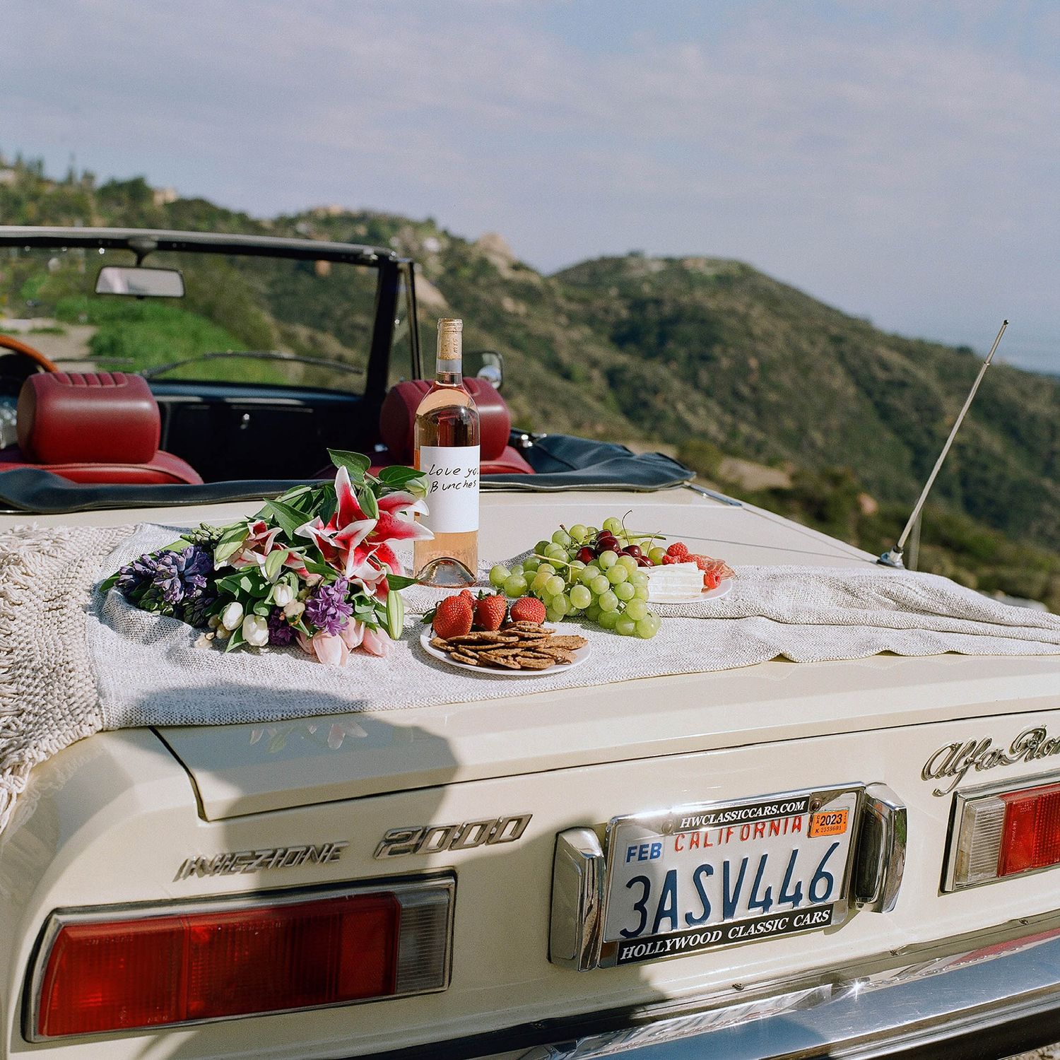 Vintage Alfa Romeo Spider convertible with California plates, topped with a romantic picnic spread of wine, grapes, and flowers.