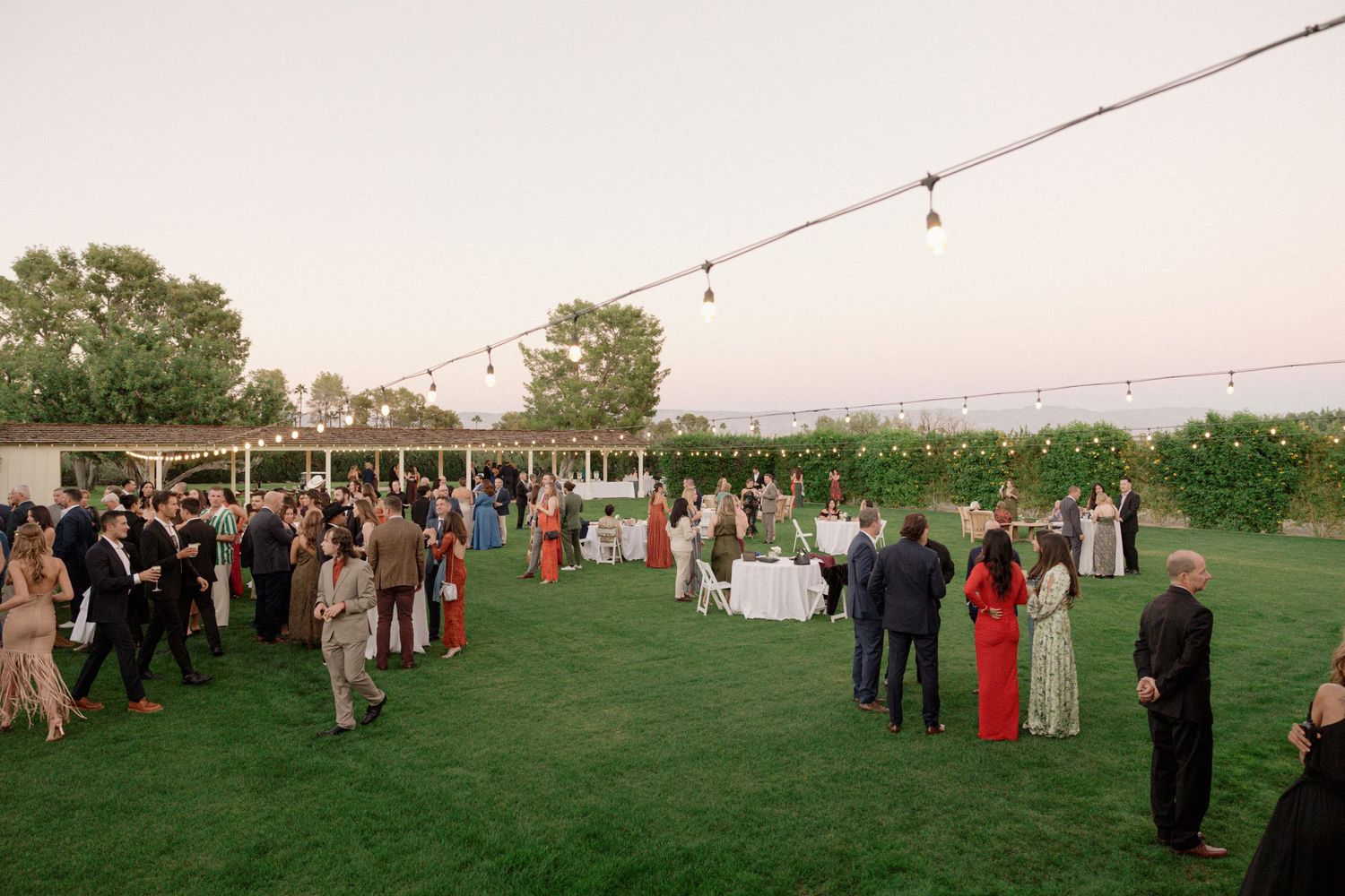 Outdoor wedding reception with string lights and guests mingling on a lawn at dusk.