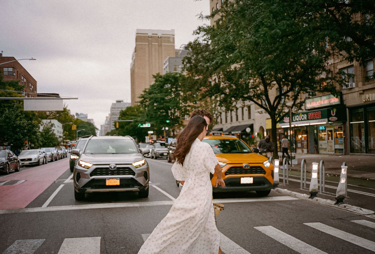 Woman in white dress crossing a zebra crossing in New York City with yellow taxi and SUV in background.