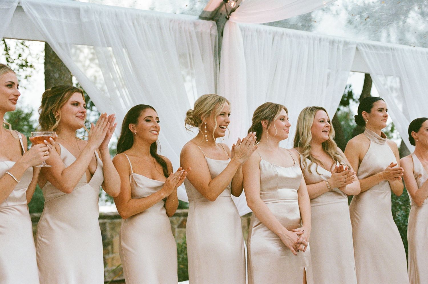 Row of bridesmaids in white dresses standing together under a white tent at an outdoor wedding.