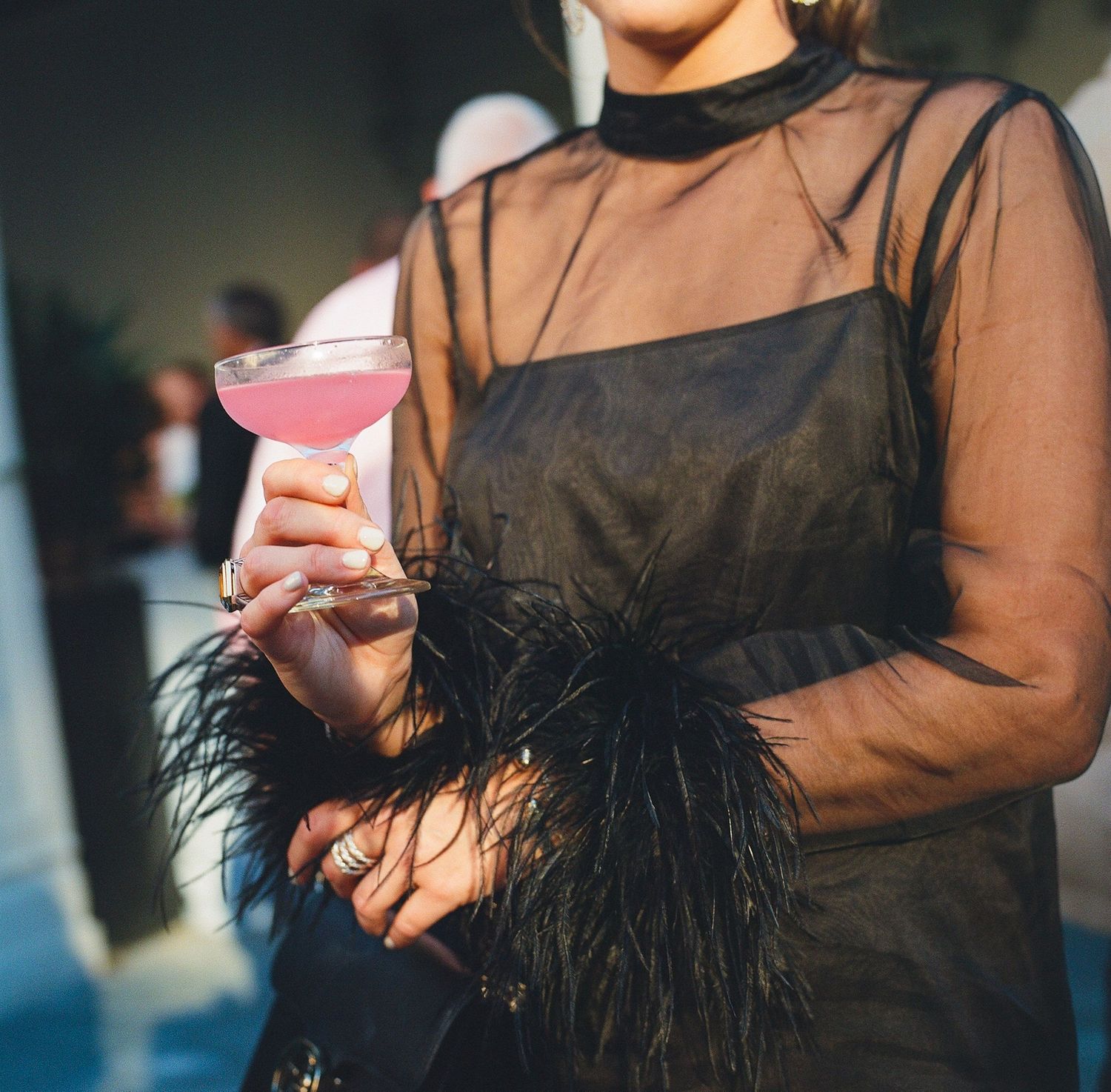 People holding pink drinks in black sheer outfits with feather details.