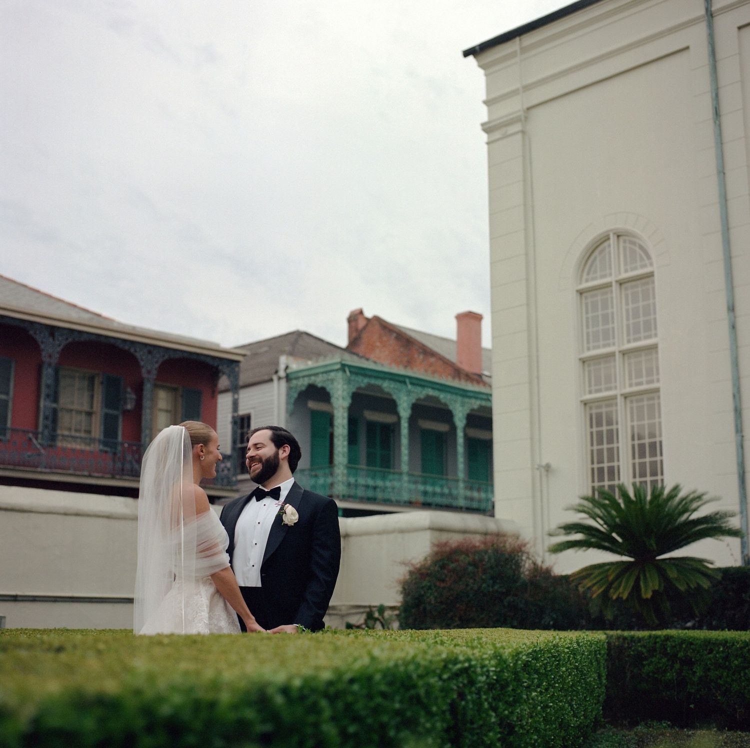 Newlyweds walk together through a formal garden with historic buildings in the background.