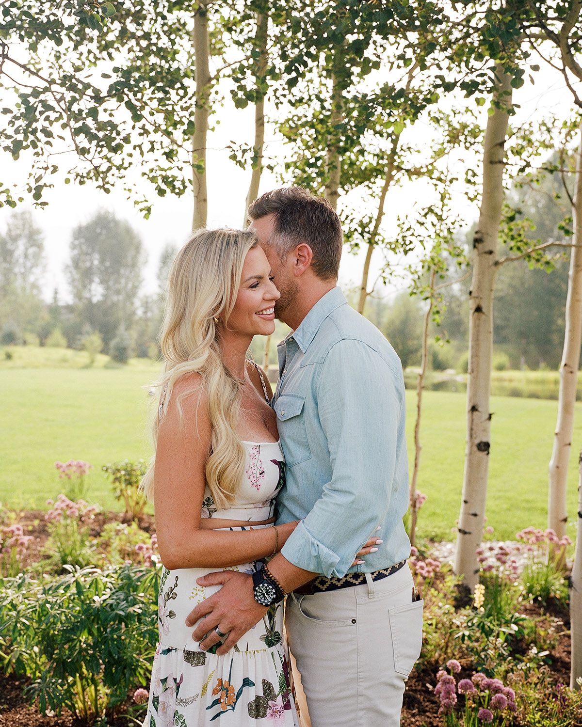 Couple embracing and smiling among aspen trees and wildflowers in a lush green outdoor setting.