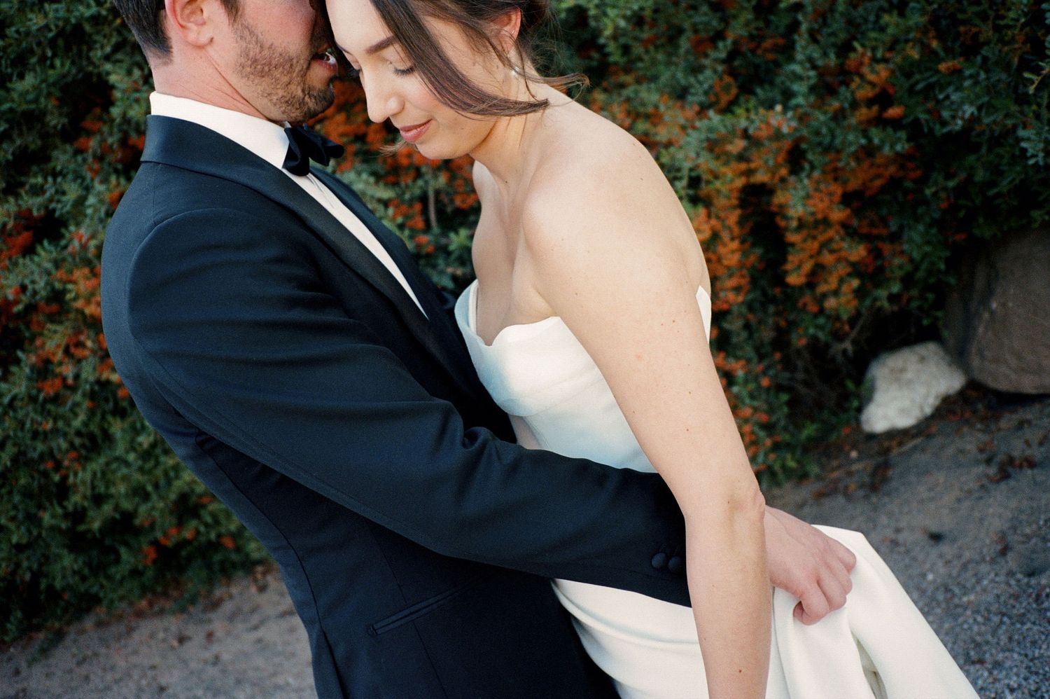 A romantic embrace between two people in formal wedding attire against a vine-covered wall.