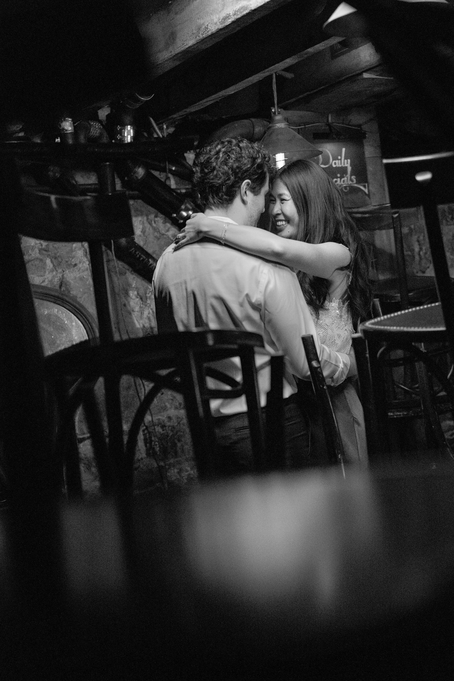Black and white photo of a couple embracing and smiling in a cozy restaurant setting.