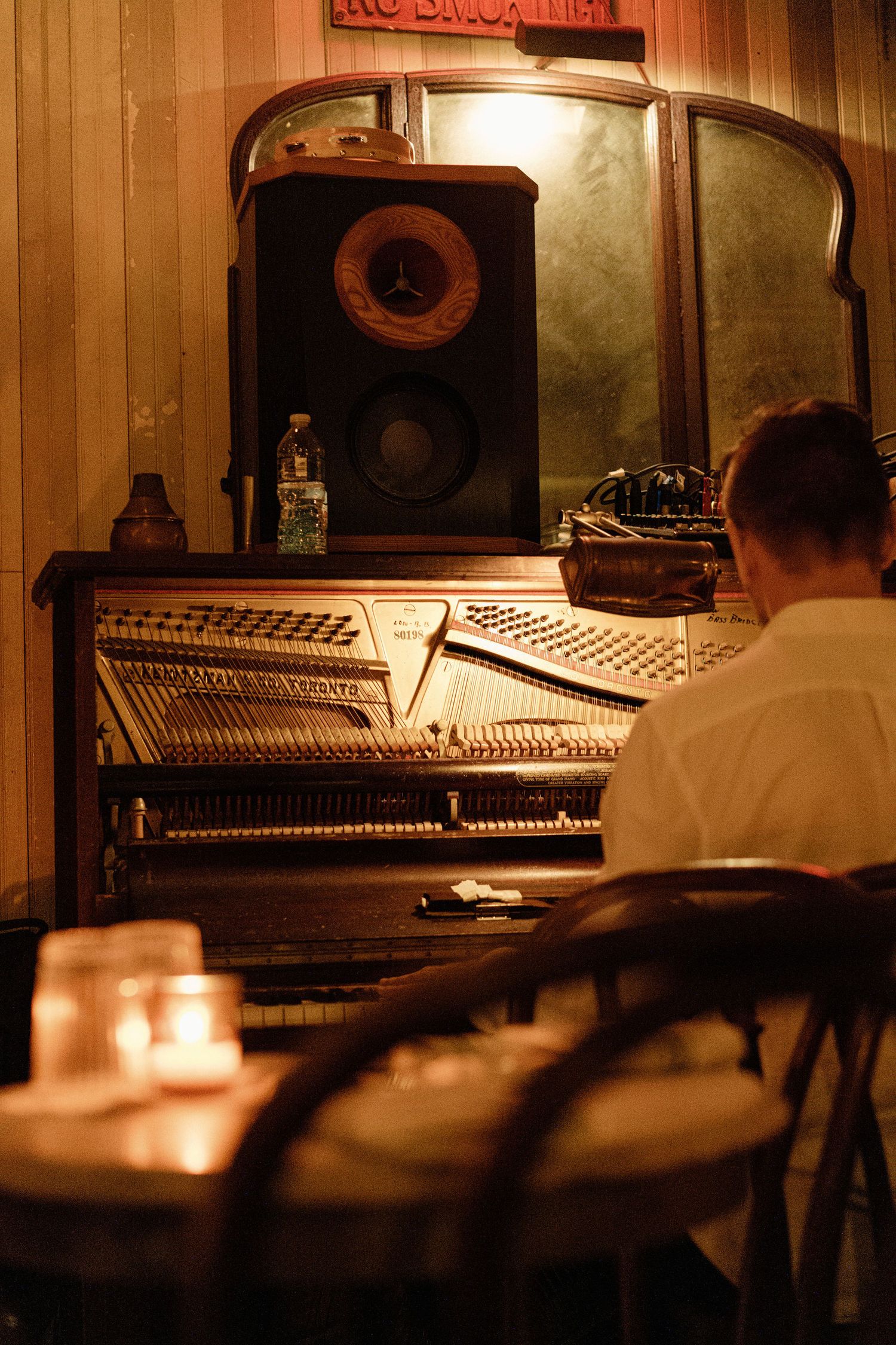 Musician playing an antique upright piano in a warmly lit, candlelit bar with a vintage speaker on top.