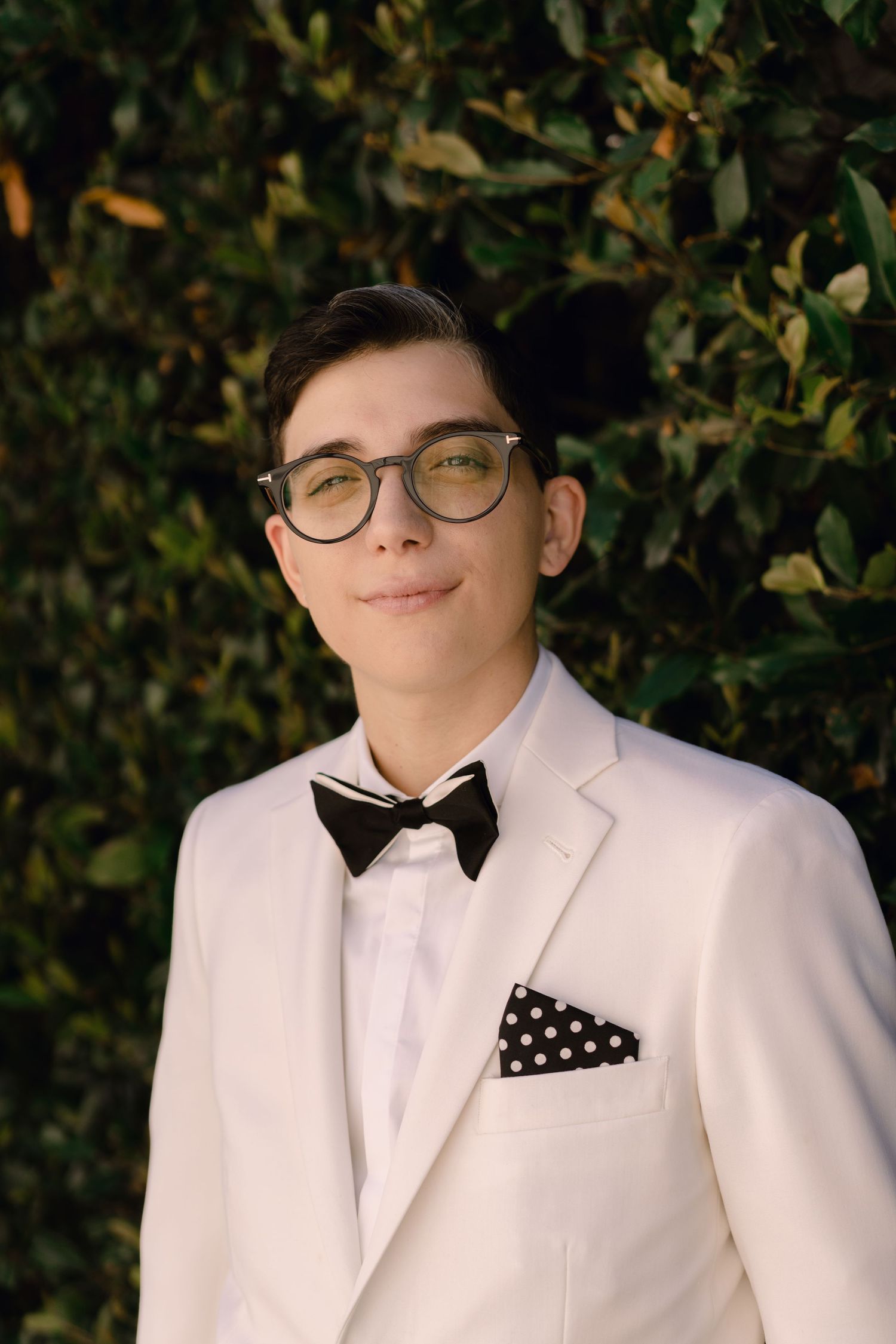 Formal portrait in white tuxedo with black bow tie and polka dot pocket square against foliage backdrop.