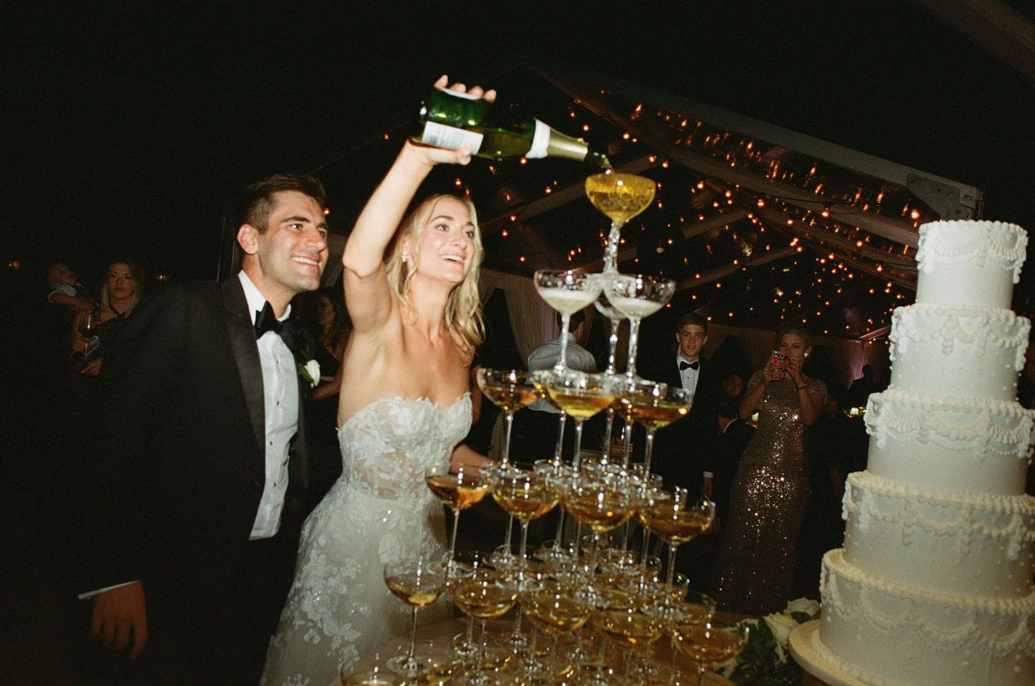Wedding reception featuring champagne tower and white tiered cake in evening lighting.