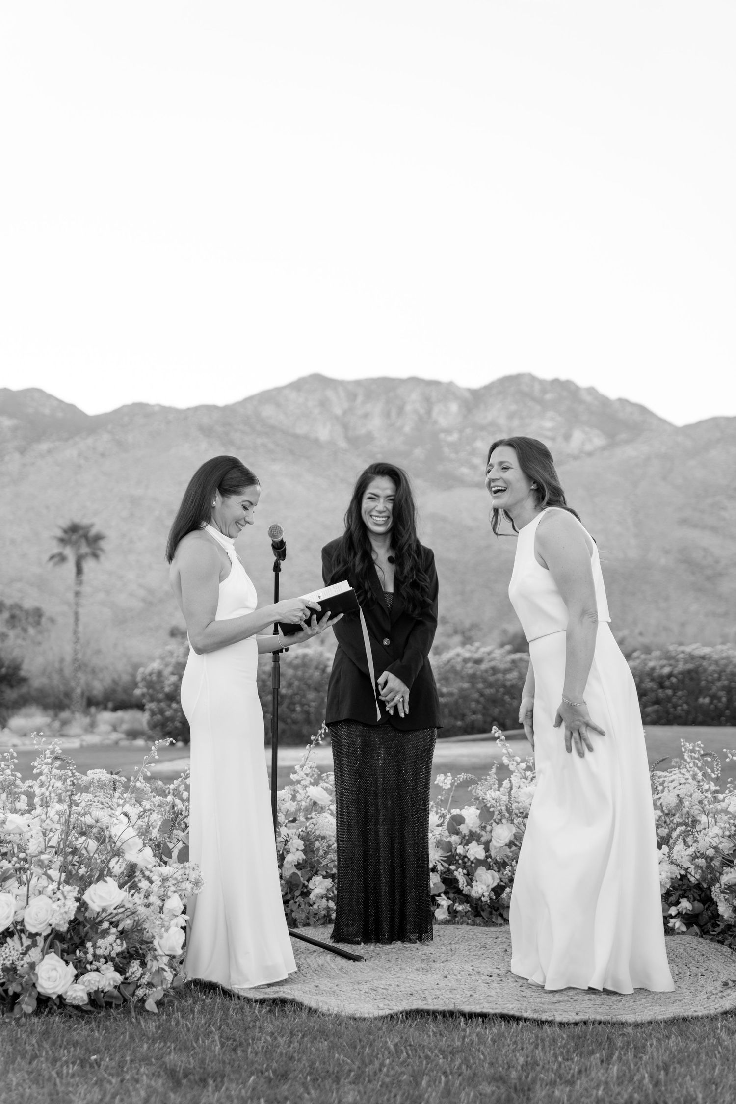 Three people in formal attire stand against mountain backdrop during wedding ceremony.