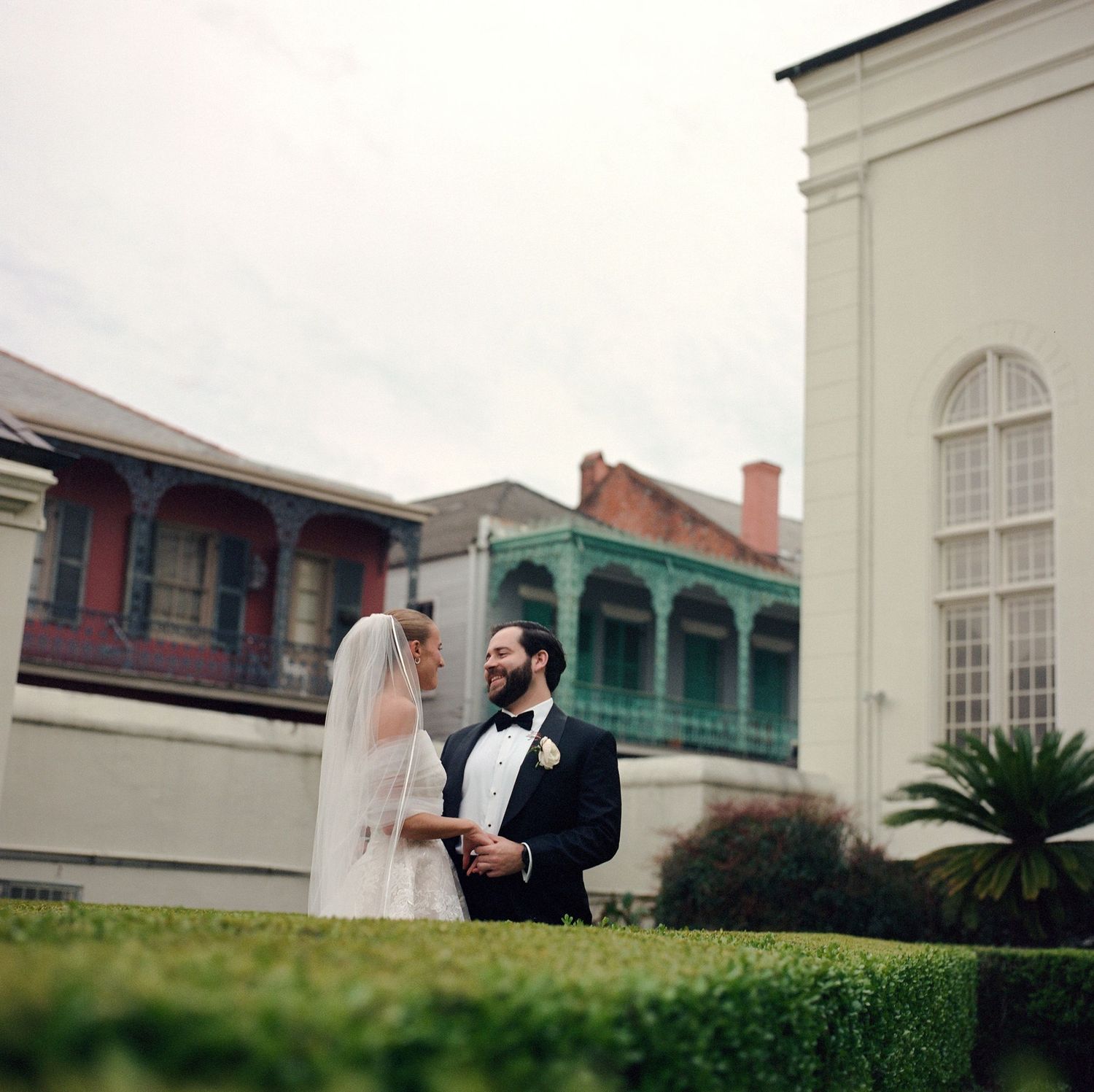 A wedding couple poses together on a scenic garden path surrounded by manicured hedges and historic architecture.