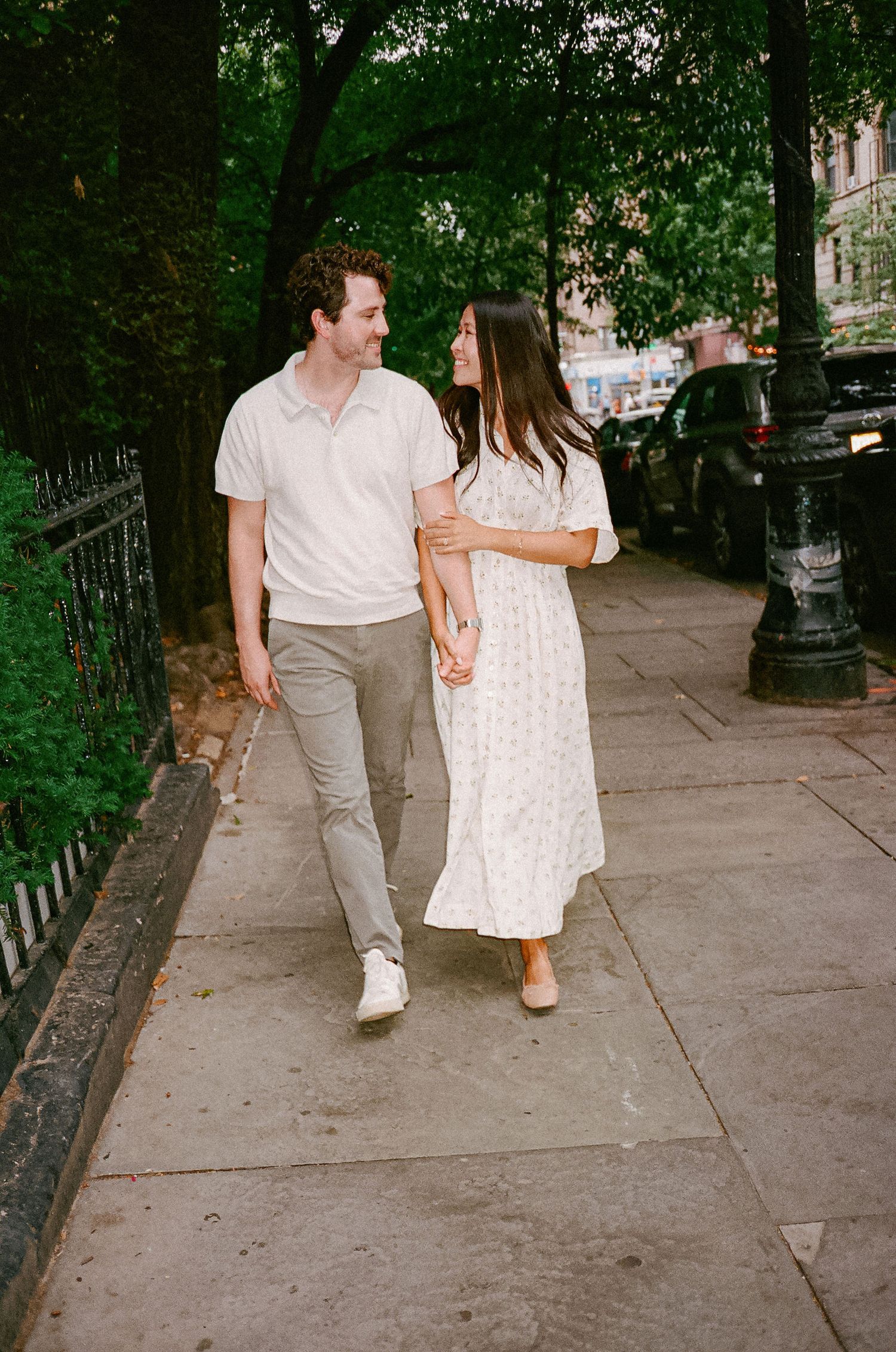 Couple walking hand in hand on a tree-lined city sidewalk, smiling at each other in casual summer outfits.