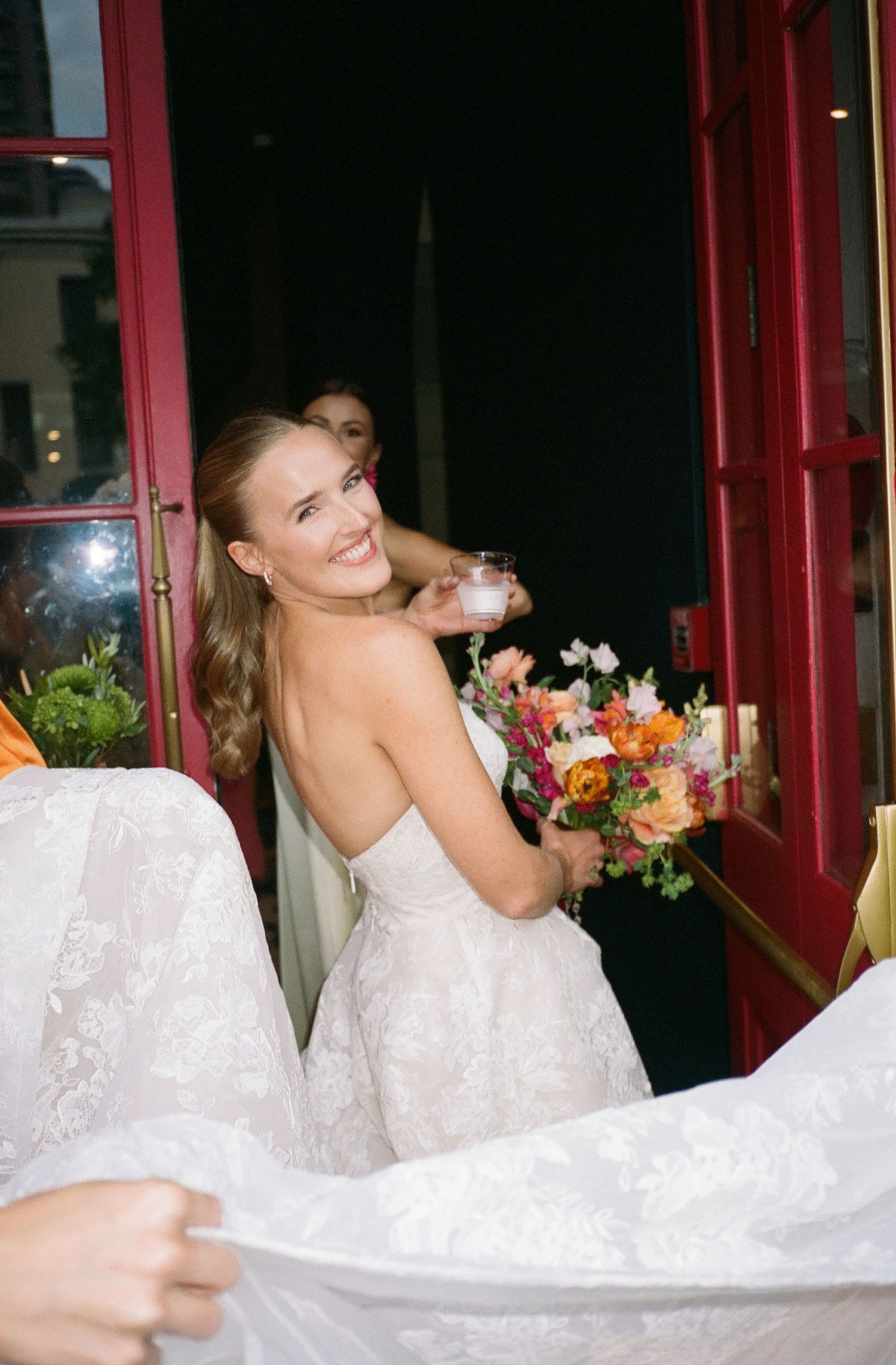 Bride in white lace gown laughs joyfully during wedding photo session by red doorway.