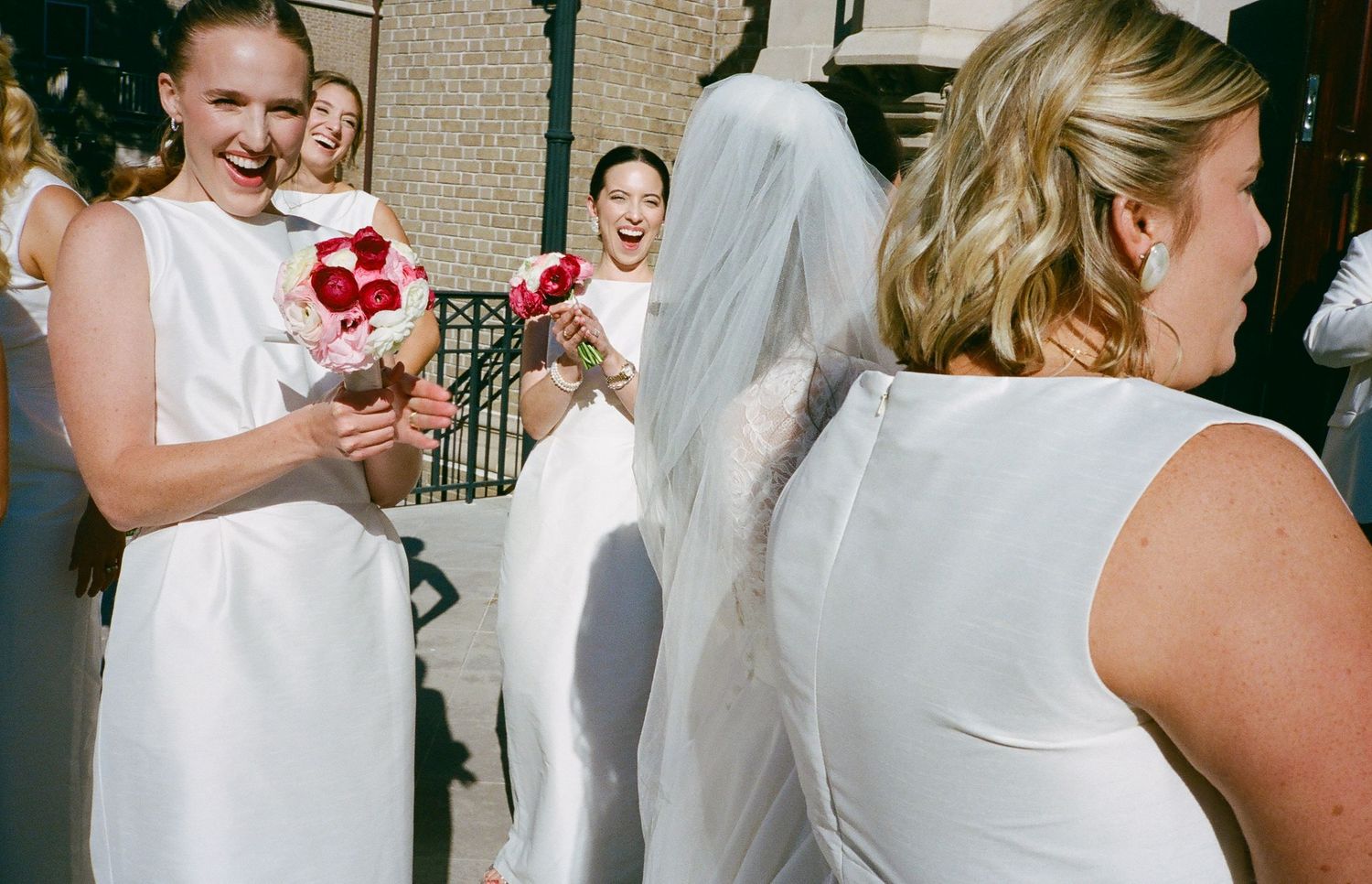 Bridesmaids in white dresses hold pink and red bouquets at an outdoor wedding celebration.