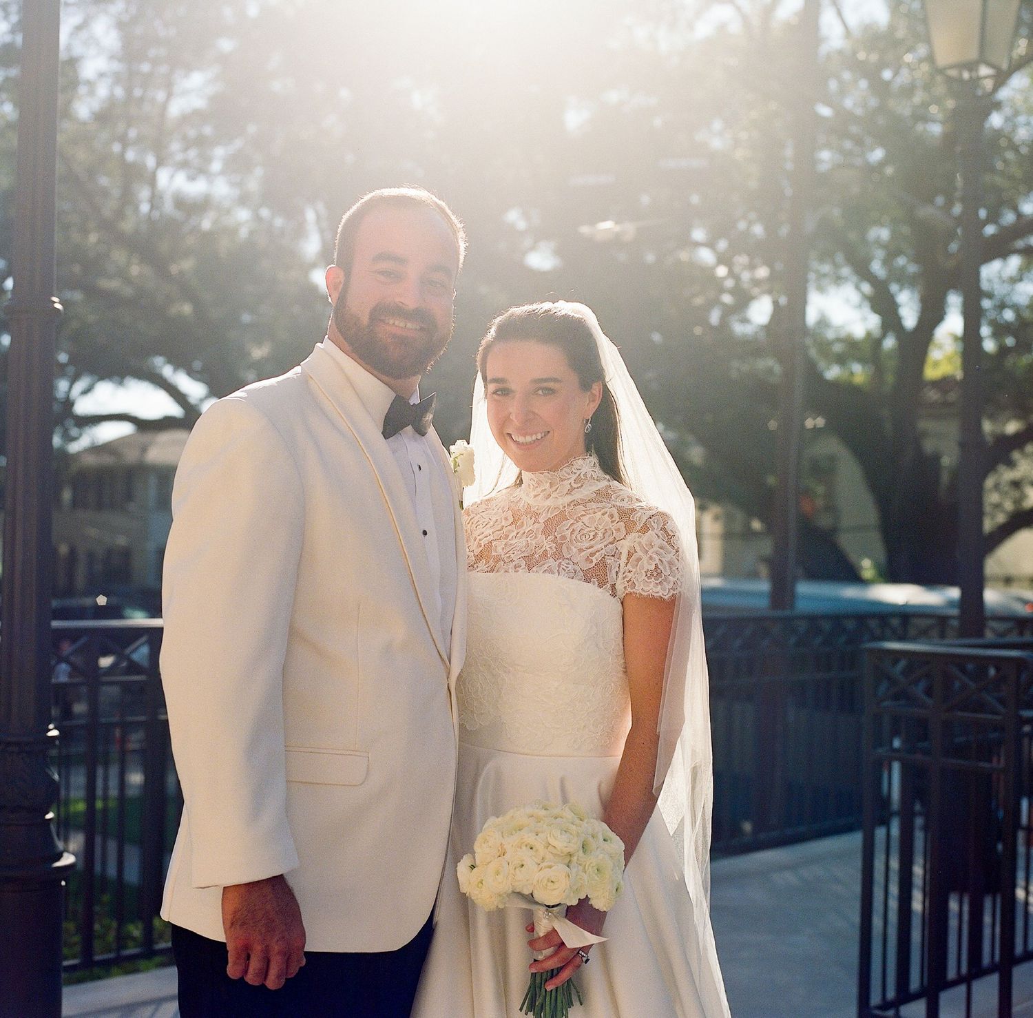 A couple poses outdoors in wedding attire with sunlight streaming behind them.