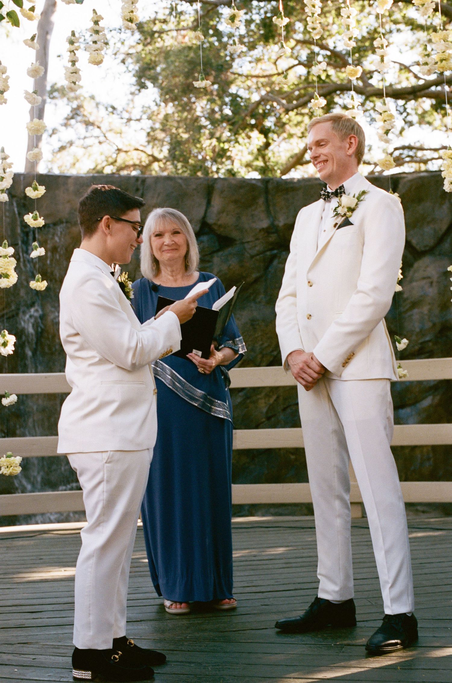 Wedding attendants in white suits and navy dresses stand on a wooden deck.