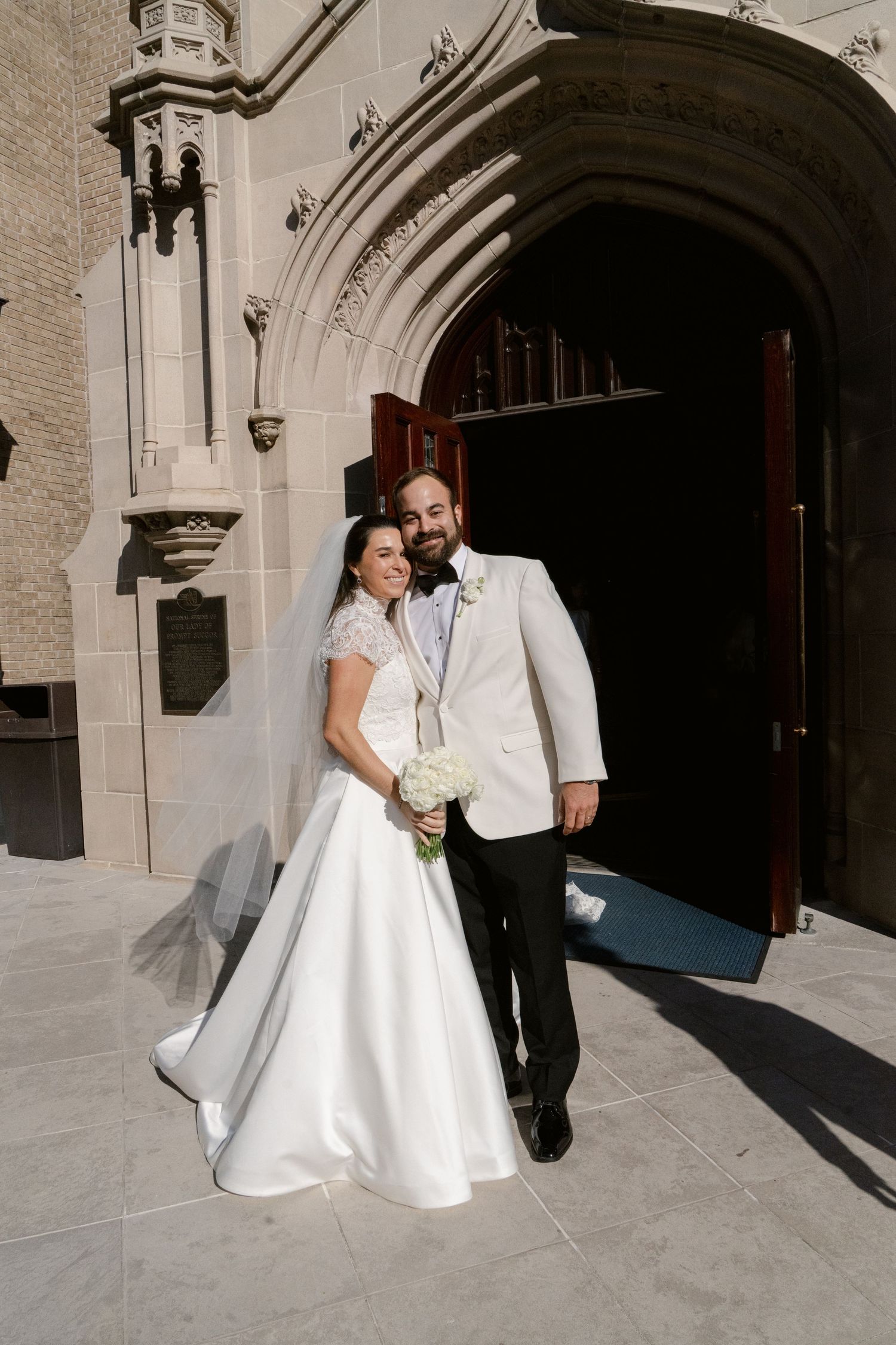 A bride in a white dress and groom in formal attire pose together against a stone church exterior.