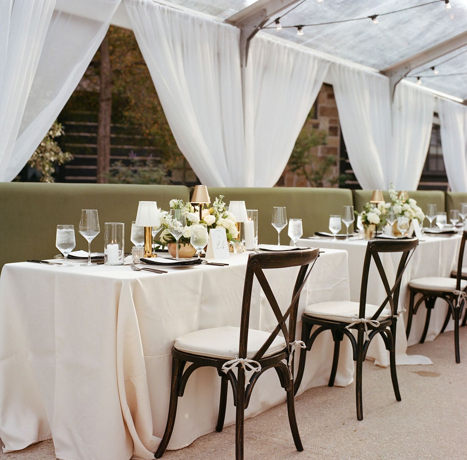 Romantic wedding reception setup with white draping, cross-back chairs and elegant table settings.